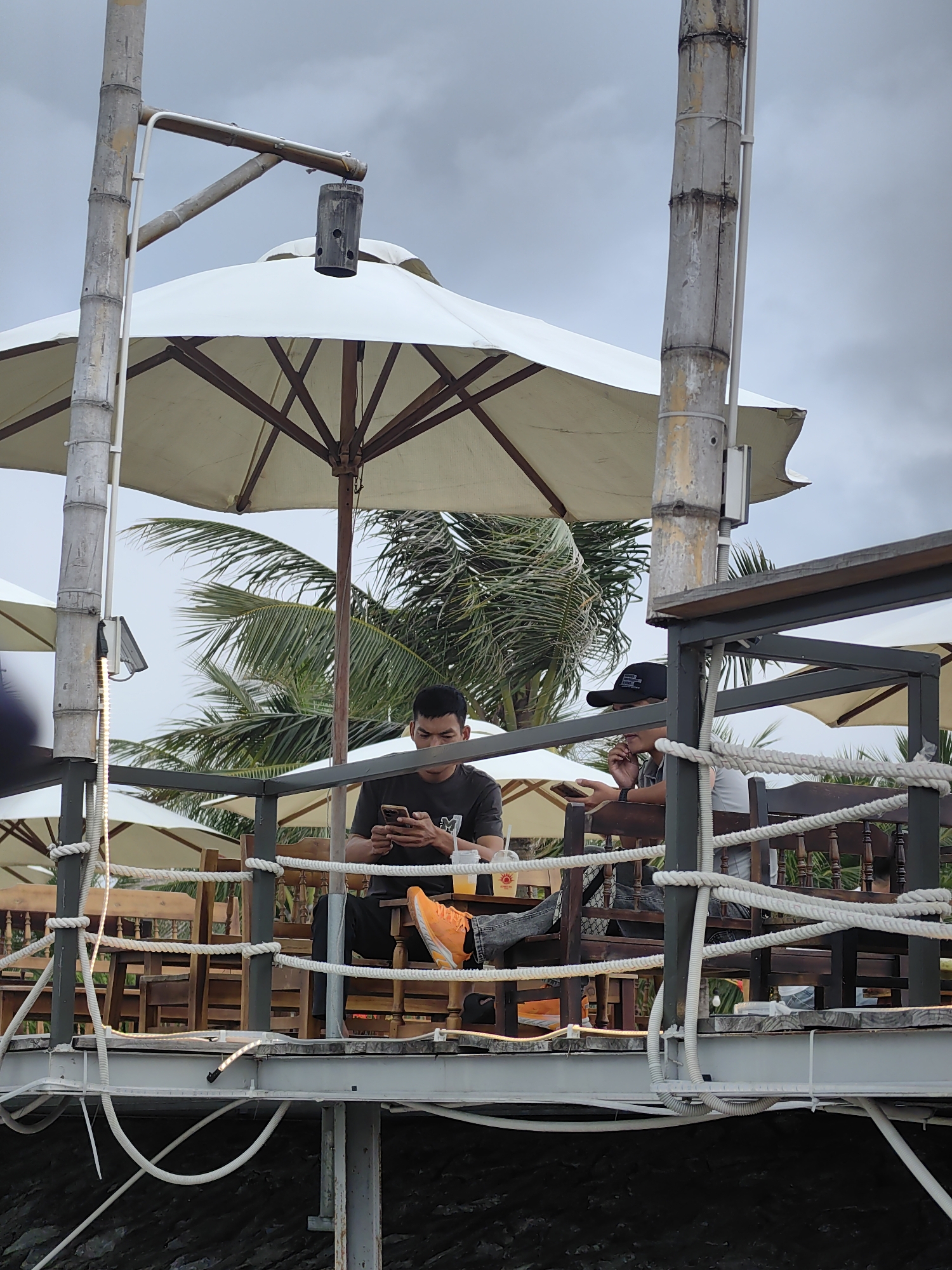 This image captures a relaxed scene at an elevated outdoor establishment, likely a cafe or restaurant, in a tropical setting. Two men are seated at wooden tables, surrounded by large, light-colored parasols providing shade.
The man on the left, wearing a dark t-shirt and dark pants, is engrossed in his smartphone, holding it with both hands and looking down. He sports bright orange sneakers that stand out against his dark clothing. The man on the right, wearing a black baseball cap and a light-colored top under a darker garment, has his hand near his mouth, appearing to be eating or drinking from a cup. He also seems to be holding a device or object with his other hand.
The structure features prominent bamboo poles, which serve as supports and parts of the railing. White nautical ropes are woven between the railings, adding a distinctive decorative and functional element. A dark, cylindrical object, possibly a speaker or lamp, hangs from a bamboo arm. Along one of the bamboo poles, a strip of LED lights is visible, and various white electrical conduits and cables run along the underside of the platform and posts, indicating functional infrastructure. In the background, lush green palm trees are visible, hinting at a warm, possibly coastal, location. The sky is overcast, suggesting a cloudy day or a diffused light environment without direct sunlight.
There is a cup on the table next to the man in orange shoes, which has an indistinct sun-like logo. No other text is clearly legible on clothing, hats, or other objects within the frame. The overall atmosphere appears casual and leisurely.