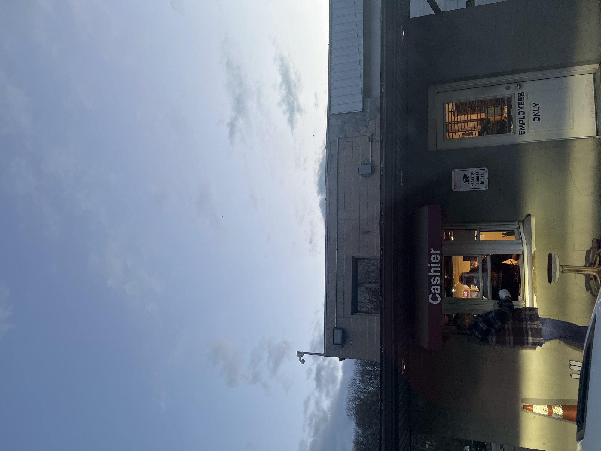 The image shows the exterior of a building in Marlboro, USA. The building has a cashier window with a red awning. A person wearing a plaid shirt and jeans is interacting with the cashier through the window. An orange traffic cone and the side of a white vehicle are visible near the cashier window. Above the cashier window is a security camera. Further up on the building, there is a door labeled "Employees Only" with a sign indicating "Security Cameras in Use." The sky is visible in the background and shows overcast conditions.