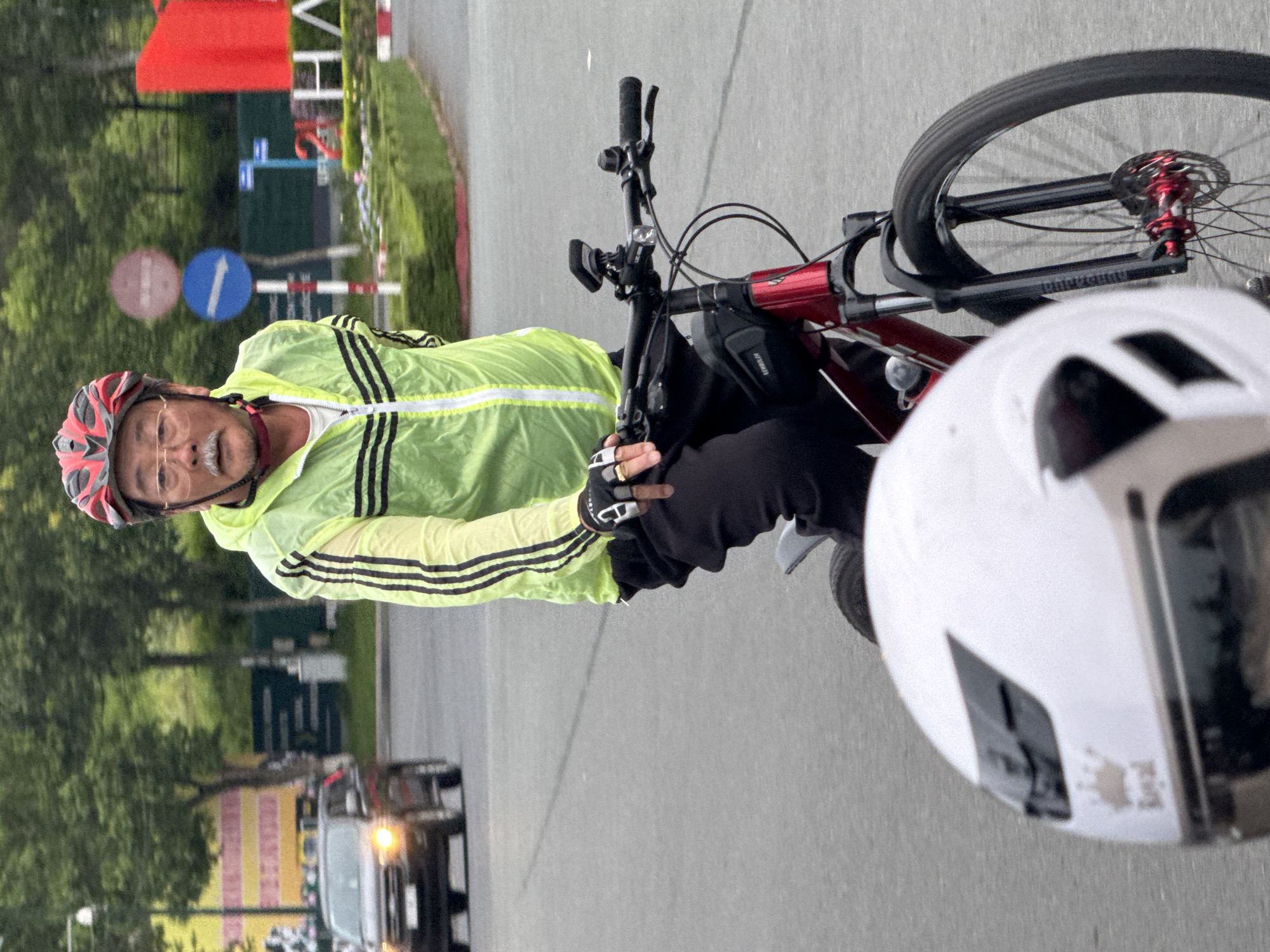 An older East Asian man wearing a red and black helmet and a neon yellow jacket with black stripes is riding a red bicycle on a paved road. He is wearing glasses, has a mustache, and is wearing black pants and black cycling gloves. He has a gold ring on his left index finger. A white helmet is in the foreground, obscuring part of the bicycle's front wheel and fork. In the background, a dark-colored car with its headlights on is driving away from the camera. Further in the background, there are trees, a red and white striped pole with two circular signs, and a large red sign with white lettering that says "HA". This scene is consistent with the urban environment of Da Lat, Vietnam, a city known for its cycling routes and scenic drives.