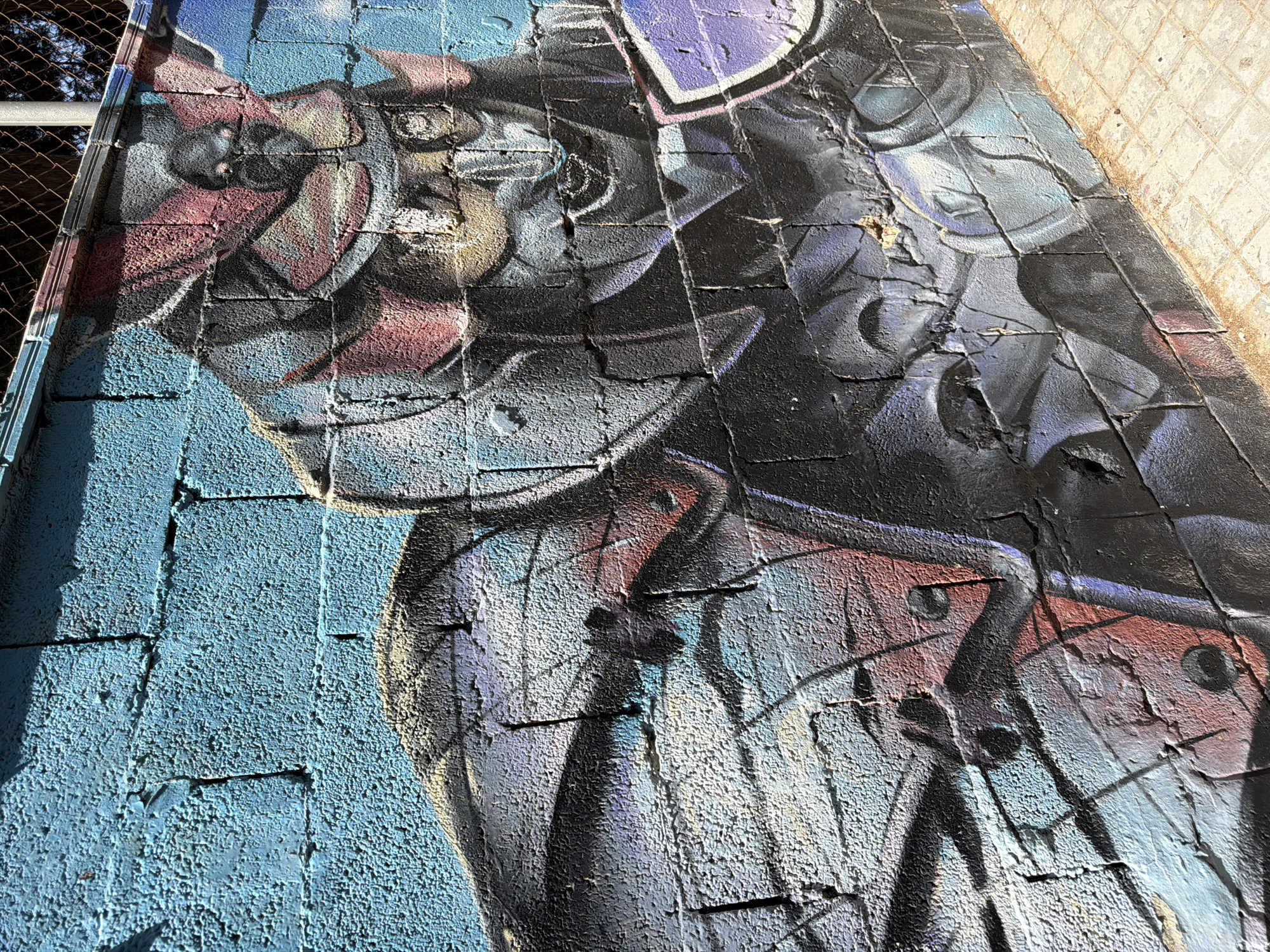 This media file is a close-up, angled shot of graffiti on a textured brick wall. The graffiti depicts a stylized, abstract figure or creature with a somewhat metallic sheen, rendered in shades of blue, purple, grey, black, and hints of red. The paint appears to be flaking and peeling in places, revealing the underlying brick.
The image is taken in bright daylight, as indicated by the strong shadows cast across the wall, particularly on the left side. The wall itself is made of rectangular bricks, with a rough, porous surface. To the left of the graffiti, a section of chain-link fence is visible, suggesting an outdoor location, possibly urban. The upper right portion of the wall transitions to a different textured material, possibly concrete or another type of masonry, laid in a tile-like pattern.
There is no visible text in the image. The graffiti itself is the primary subject, with no discernible activity or event taking place beyond its artistic rendering. The overall impression is one of urban art on a weathered surface, captured in stark sunlight.