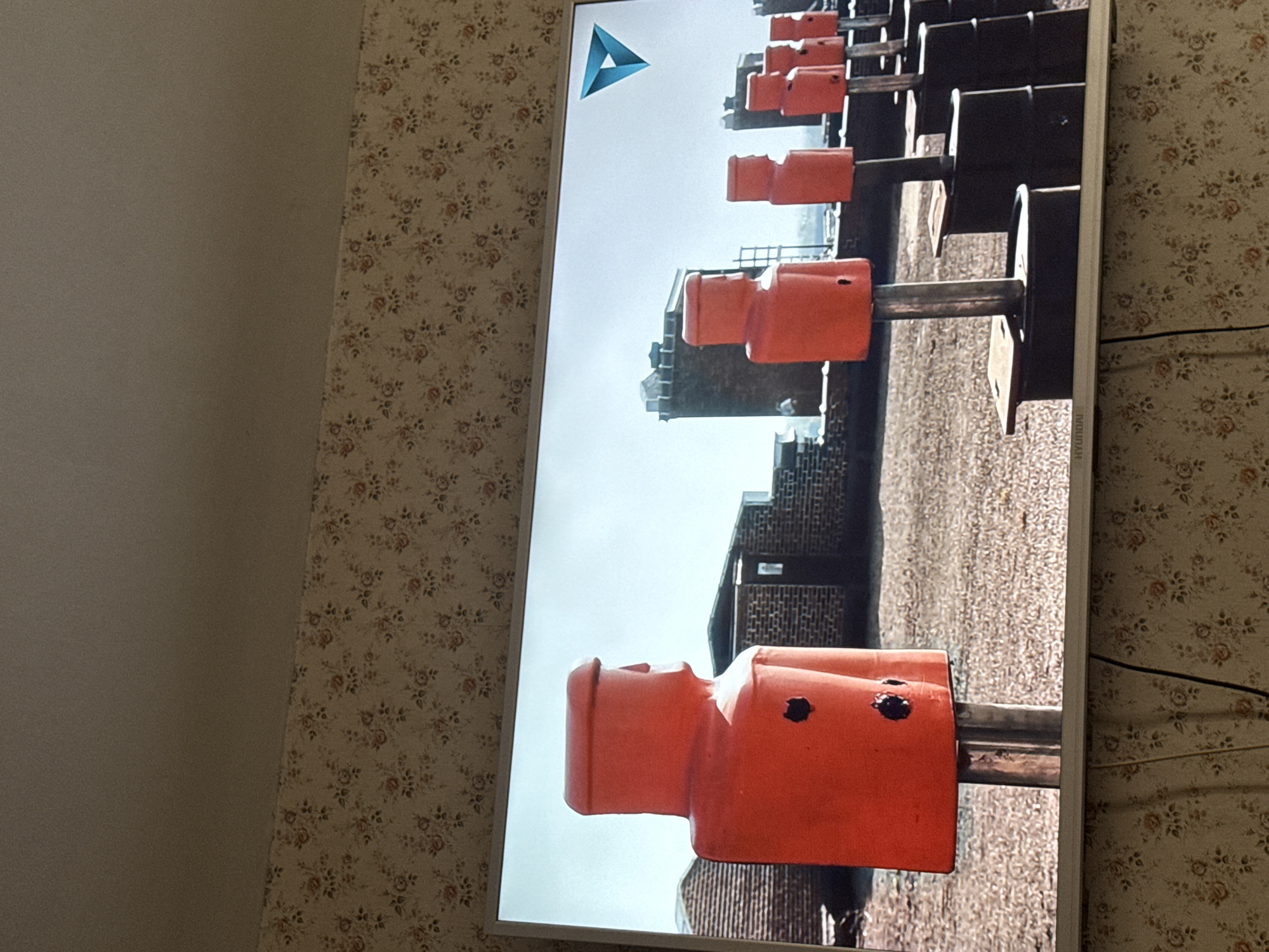 A television screen displays a scene of a foosball table, likely in Mytishchi, Russia, based on the presence of the "Delta" logo (a common developer in the Moscow area) at the upper left corner of the screen and architectural characteristics in the background. The camera angle is low, looking up at the orange foosball players attached to rods, revealing a cityscape in the background that includes residential or office buildings. The walls surrounding the television are covered with wallpaper featuring a floral pattern.