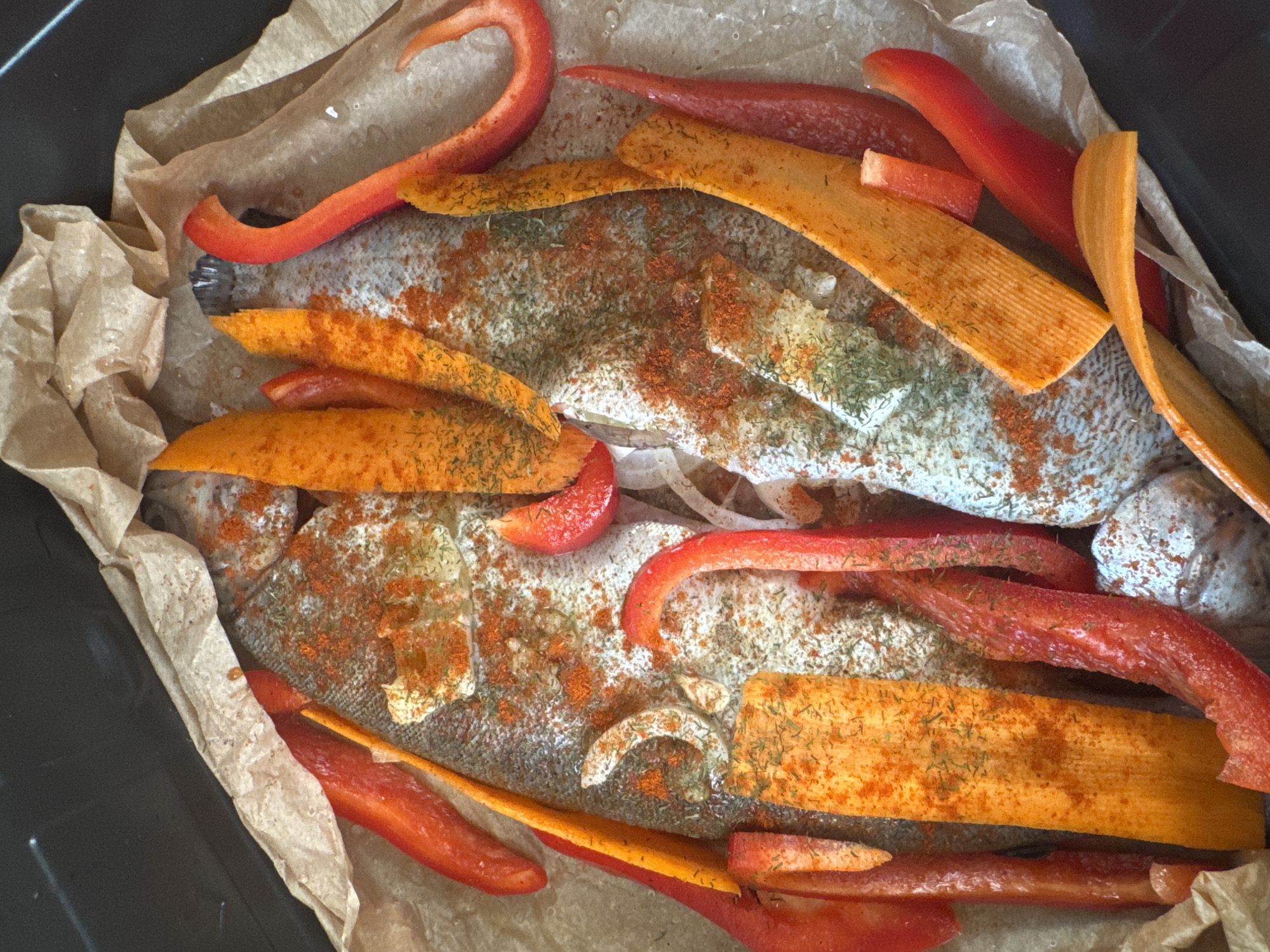 The image shows two whole fish, likely trout, seasoned with paprika and herbs, arranged on parchment paper in a baking tray. Sliced red bell peppers and carrots are placed around and on top of the fish. The overall impression is of a meal being prepared for cooking. The image does not contain any visual cues or information that would allow for the determination of its location within the city of Katowice, Poland, or any other specific geographical location.