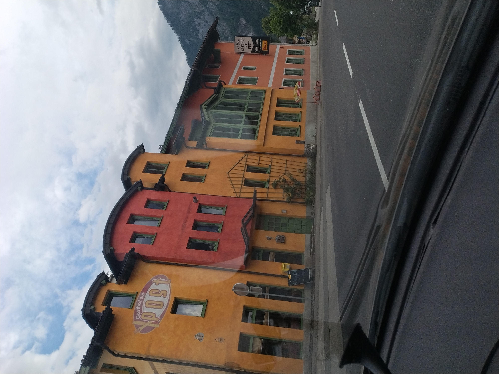 In St. Agatha, Austria, a two-story building with an orange and red facade, identified as "Gasthof zur Post," is visible from the perspective of a vehicle. The building features multiple windows, some with green frames. A sign above the entrance reads "Gasthof zur Post" with an emblem of a post horn. To the right of the building, another sign displays "Gasthof zur Post" and a website address. The scene depicts a street with a crosswalk and markings, suggesting a public area. Mountains are visible in the background. The sky is cloudy.