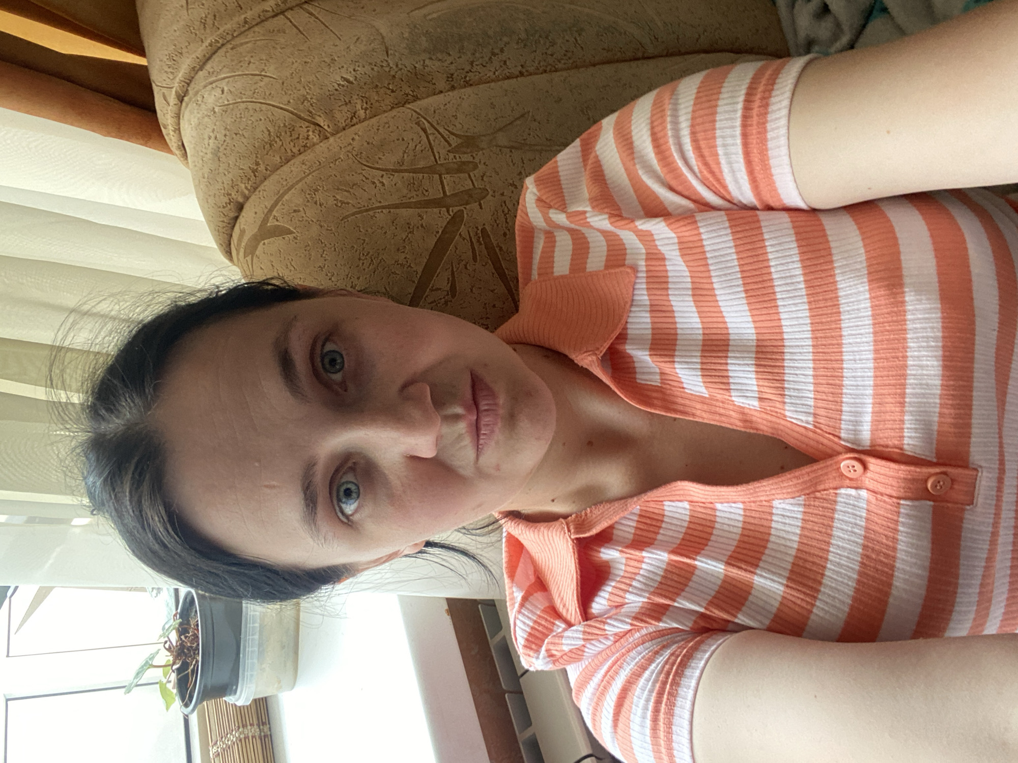 The image shows a woman lying on her side on a couch. She is wearing a striped orange and white shirt with a collar. Her dark hair is pulled back. Her eyes are open and she is looking towards the camera. The couch appears to be upholstered in a textured, beige fabric. A window with sheer white curtains is visible in the background, along with a potted plant. The lighting suggests it is daytime.