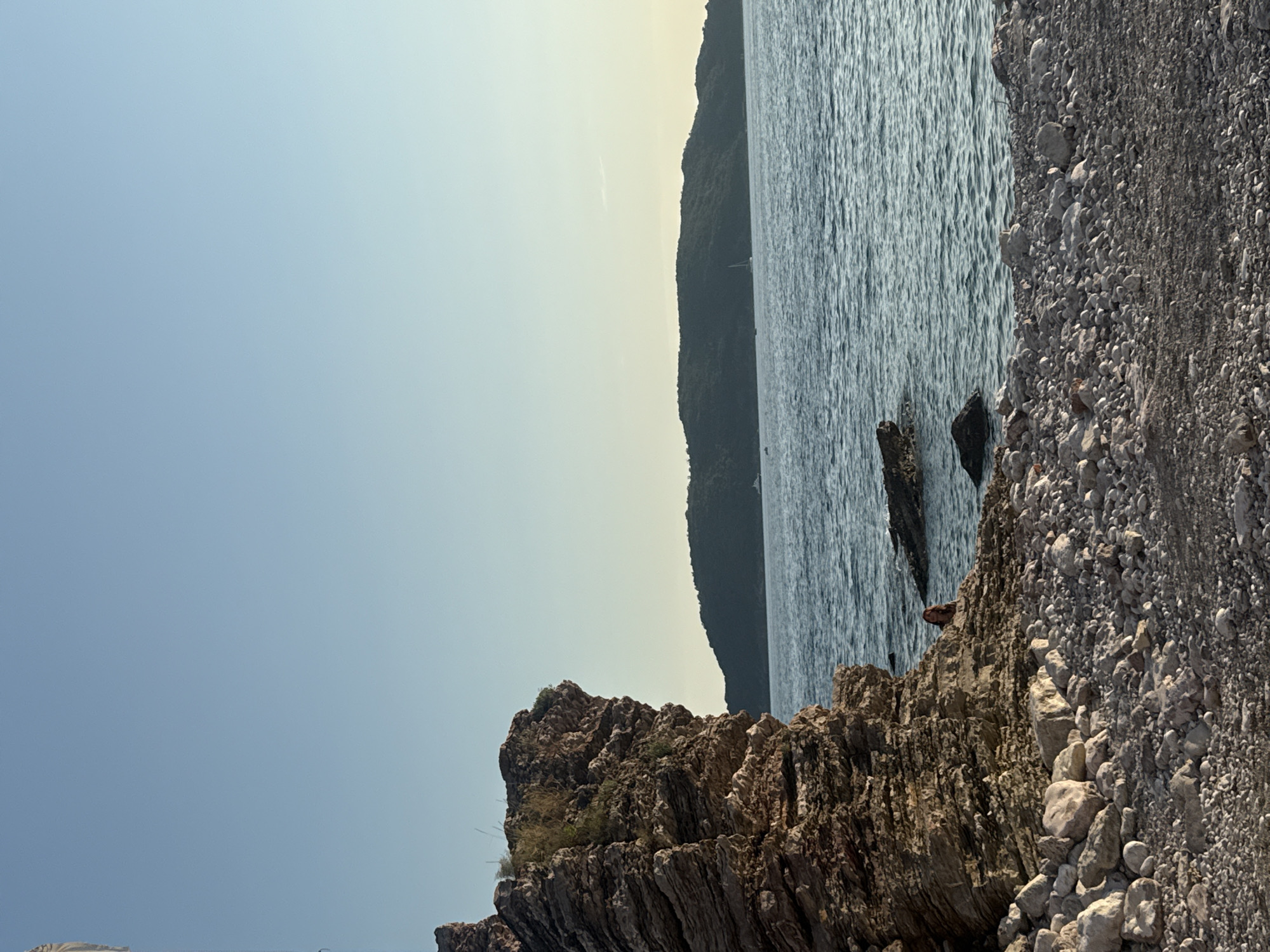 A landscape photograph captures a rocky coastline in Rafailovići, Montenegro, under a clear, light blue sky. The foreground is dominated by a pebble beach strewn with various sizes of grey and white stones. To the left, a large, layered rock formation with a rugged texture rises from the beach. A single person with reddish-brown hair is seated on a rock formation near the water's edge, partially obscured by the cliff. Beyond the shore, the calm, blue-green Adriatic Sea stretches out, dotted with a few distant boats and a sailboat. Across the water, a verdant, hilly island creates a natural backdrop, its silhouette softened by the distance and the warm, late afternoon light. The overall impression is one of peaceful, natural beauty.