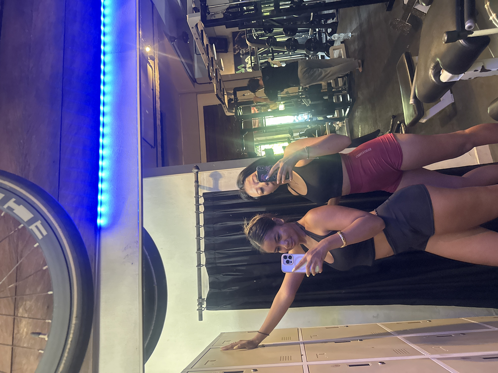 This is a full-body, mirror selfie taken at a gym in Denpasar, Indonesia. Two young women are posing in athletic wear. The woman on the left, facing the camera, wears a black sports bra and black shorts, and is holding a white smartphone. The woman on the right, also facing the camera, wears a black sports bra and maroon shorts, and is holding a smartphone. In the background, other gym equipment and a person exercising can be seen. The environment appears to be a gym with lockers on the left, a black curtain behind the women, and various weight machines and benches in the background. The lighting includes blue LED strip lights overhead.