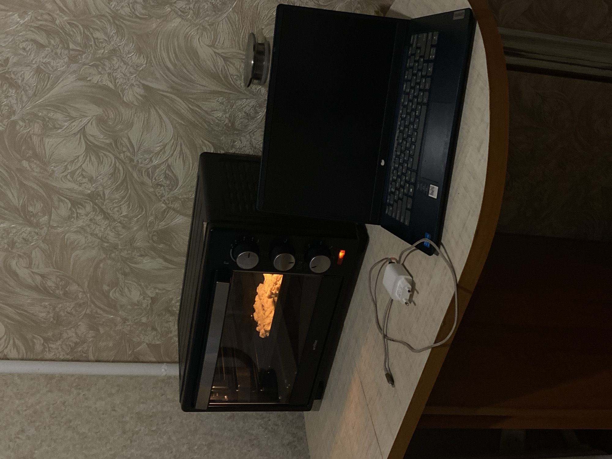 The image depicts a cluttered countertop scene in what appears to be a kitchen or dining area. Dominating the image is a black countertop oven with bread or another light-colored food item baking inside. The oven door is slightly ajar, revealing the interior contents. A black laptop is positioned to the right of the oven, partially obscuring the background wall. A power adapter and cable are connected to the laptop and draped on the countertop near the oven. The background wall features a light-colored, ornate floral pattern. The countertop itself is light beige. The location is in the city of Chaikovsky, Russia.