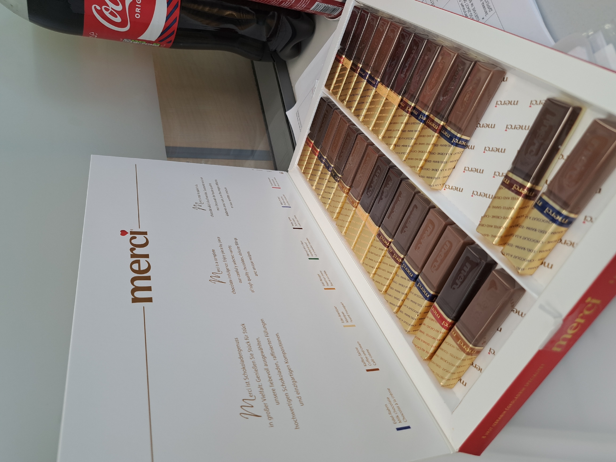 A package of Merci chocolates lies open on a white surface. The chocolates, individually wrapped in gold foil, are arranged in rows within the package. A bottle of Coca-Cola stands in the background. There are documents with Bulgarian text on them. The image appears to be taken indoors in Sofia, Bulgaria.