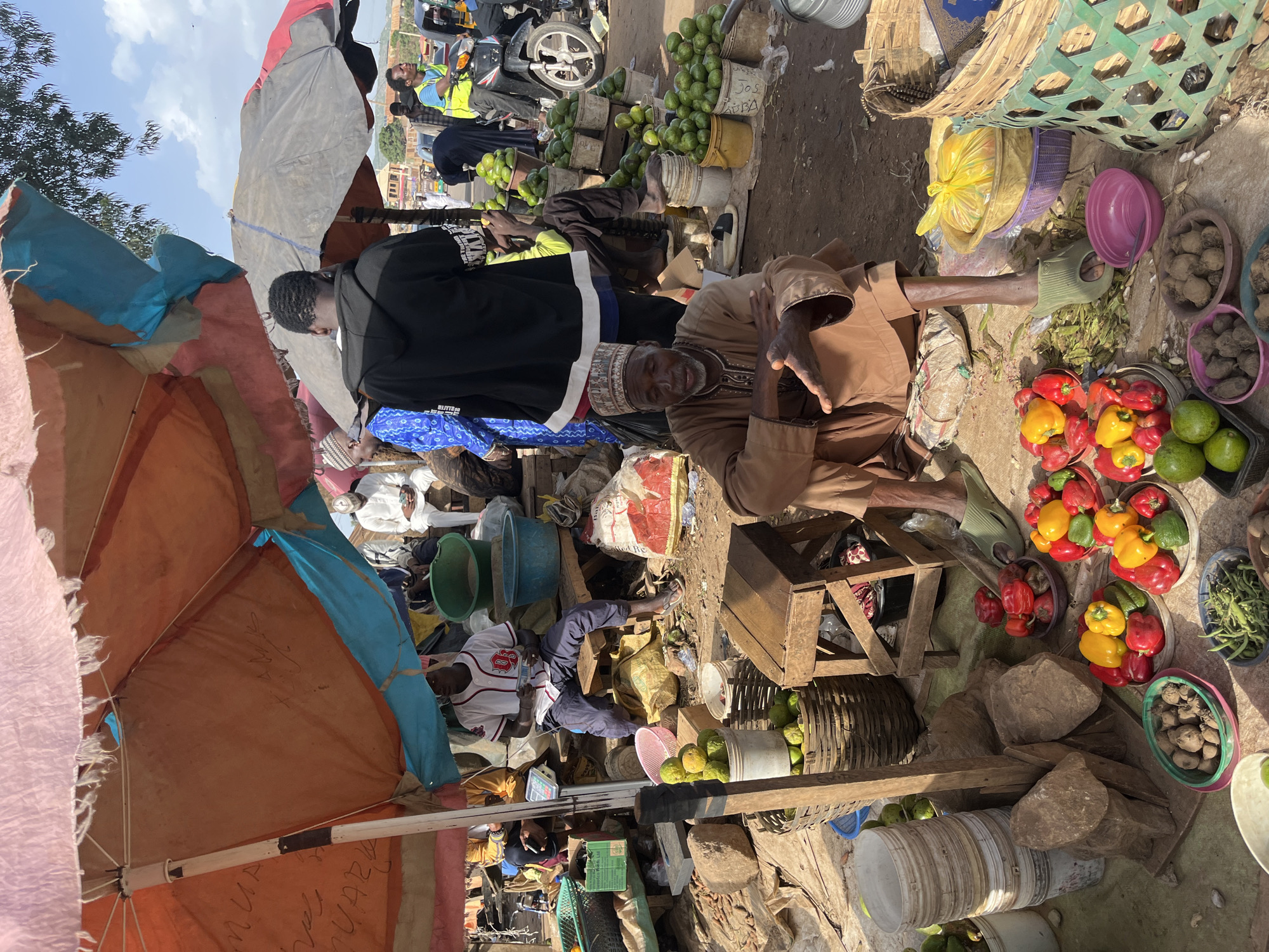 This media file captures a bustling outdoor market scene in Rafin Par, Nigeria. The image is dominated by stalls displaying a vibrant array of fresh produce, including piles of green avocados, red and yellow bell peppers, and other unidentifiable vegetables. Several people are engaged in market activities. In the foreground, an older man in traditional attire sits with his hands extended, appearing to be in conversation or making a transaction. Behind him, a younger person in a black hoodie is also present. Other individuals are visible in the background, some tending to their goods, others possibly customers or passersby.
The setting is an open-air market with makeshift stalls constructed from wood and fabric. Tarps and umbrellas provide shade, their bright colors contrasting with the dusty ground. Woven baskets and plastic containers are used to hold and display the produce. In the distance, hints of motorized transport suggest a busy thoroughfare nearby, and the presence of a motorcycle with a rider wearing a high-visibility vest further emphasizes the active nature of the location.
The time of day appears to be daytime, given the natural lighting and shadows. The weather seems to be warm, typical of a sunny day in Nigeria. The overall atmosphere is one of lively commerce and community interaction. Some of the objects in the scene include buckets, crates, and various food items. There are no overtly visible emotions expressed by the individuals, but the activity suggests a normal day of trade. While specific text is not clearly legible, some writing appears on the tarps, likely related to vendors or product names, but it is too indistinct to decipher.