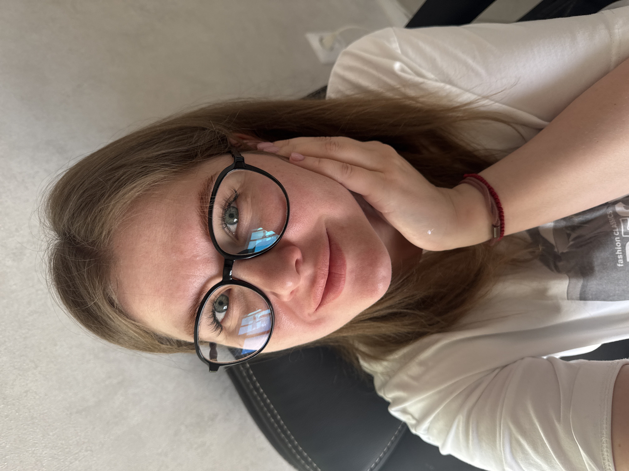 A woman with light brown hair and fair skin is posing for a selfie, sitting in what appears to be an office chair. She is wearing black, round-framed glasses and a white t-shirt. Her left hand is touching her face near her cheek. On her wrist is a red string bracelet. The background includes a light-colored wall and part of a plug. The photograph appears to have been taken indoors, likely during the daytime. There is no definitive information in the image to confirm the location is Gornyi (Dvurechenskoe S/P), Russia.