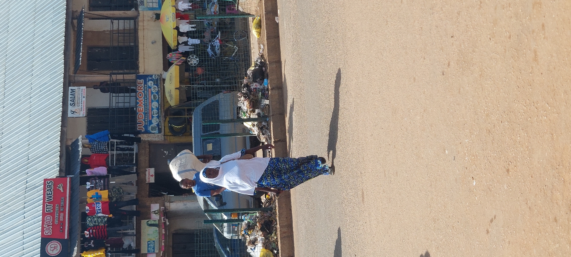 In a street scene of Unknown, Unknown, a woman wearing a white headscarf and a blue and white patterned skirt walks on the right side of a paved road. She carries a large, white bag on her head. Behind her, a man in a blue t-shirt stands near a gray van. The background features buildings with awnings, shops displaying clothing, and a wire fence enclosing an area with scattered trash and umbrellas. The road surface is a sandy brown color, with shadows of people and objects cast on it.