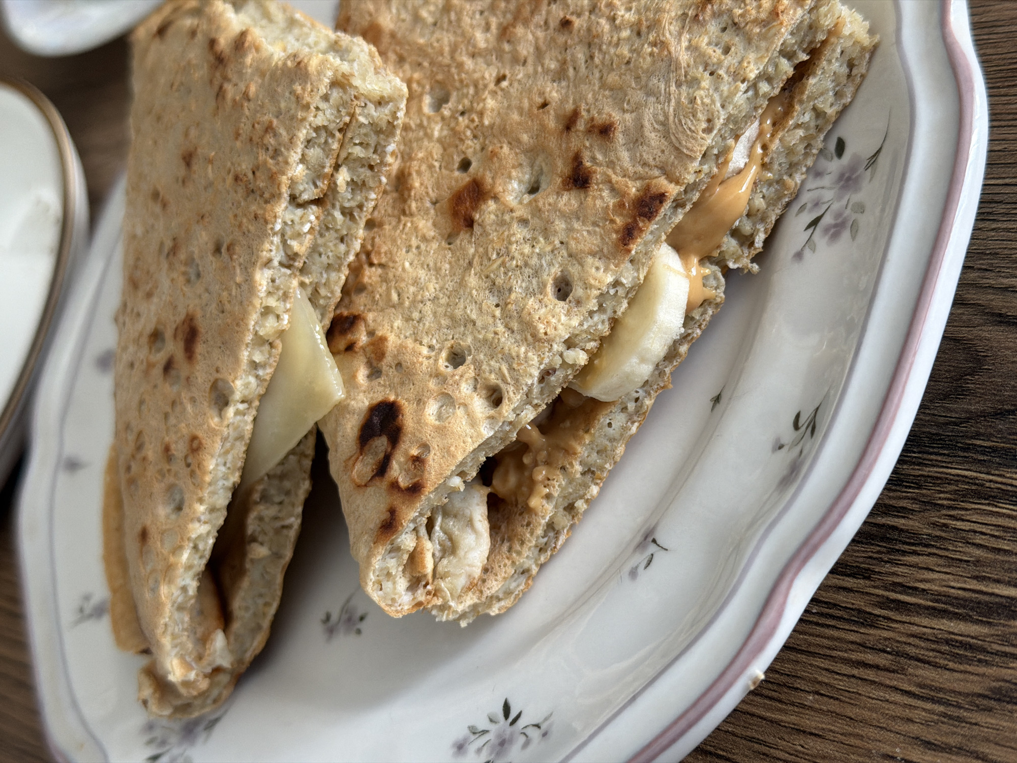 The image shows a close-up of a plate with a sandwich cut into two halves. The sandwich appears to be made with whole wheat or grain bread, and it contains a filling of peanut butter and sliced bananas. The plate is white with a floral pattern and a pink rim. The surface beneath the plate appears to be a dark wooden tabletop. There is no information on this photo to suggest a location in Perm, Russia.