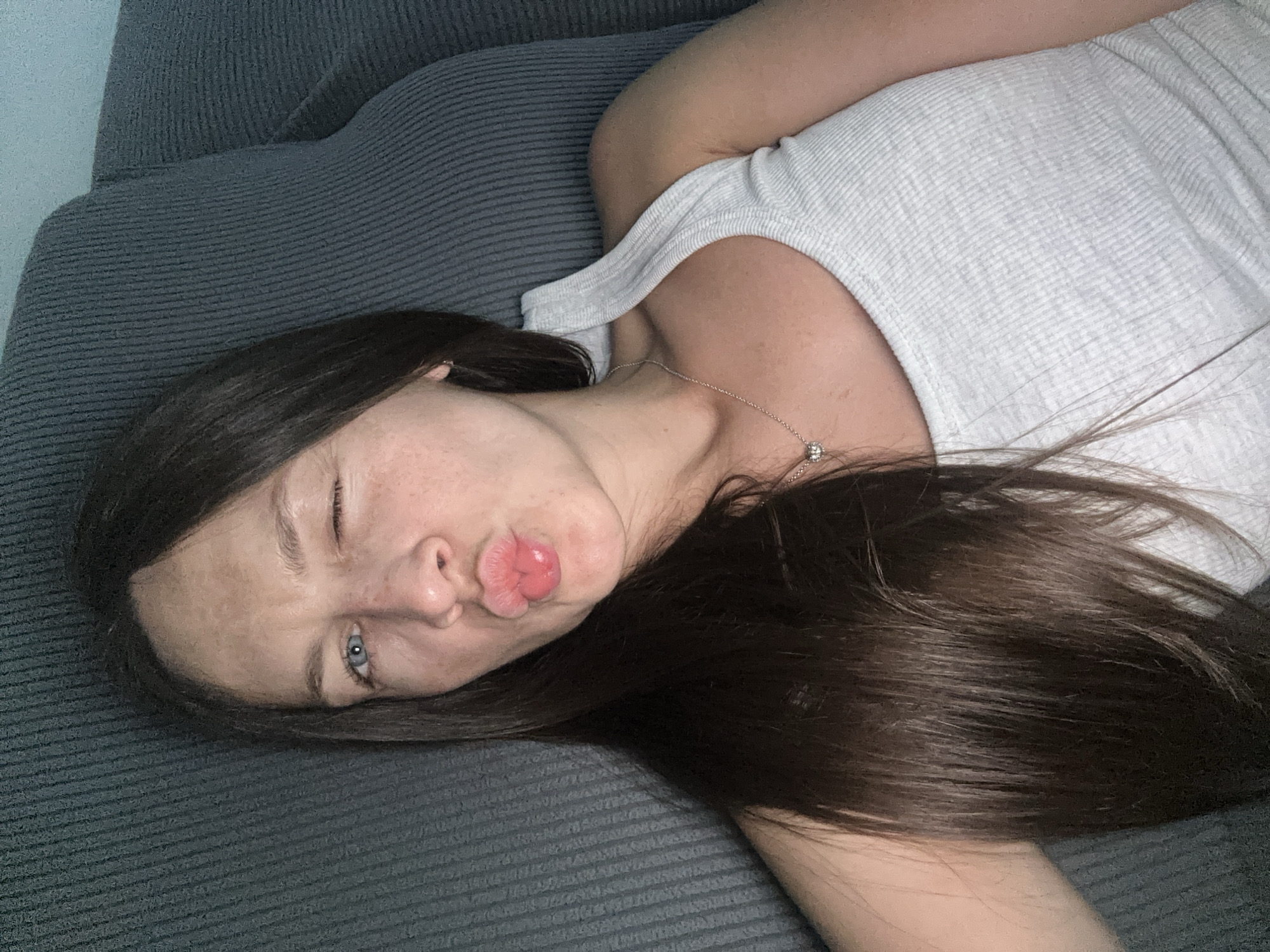 A medium shot shows a woman with long, dark brown hair lying on a gray sofa. She is looking at the camera with one eye open and her lips puckered in a kiss, giving a playful expression. She is wearing a light gray ribbed tank top and a silver necklace with a small, sparkling pendant. The sofa has a textured fabric, and a portion of what appears to be a white pillow or blanket is visible to the right. The lighting is dim, creating shadows and highlighting the woman's face and chest. The scene is indoors, and the background is dominated by the dark gray of the sofa. The location is stated as being in Donji Pobori, Montenegro.