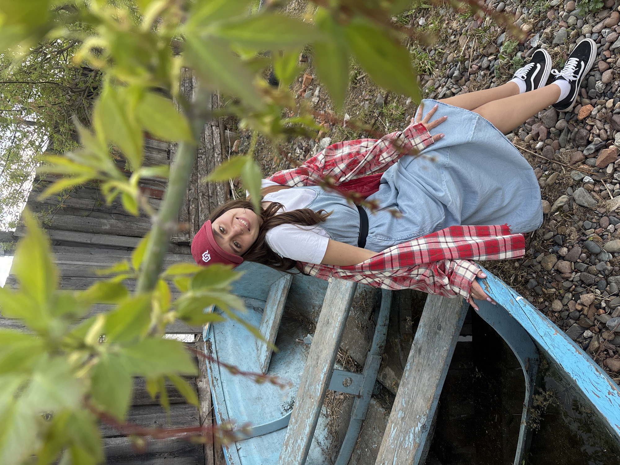 A medium shot, taken outdoors in what seems to be a rural or natural setting, presents a young woman lying in an old, light-blue boat. She is positioned lengthwise in the boat, with her head near the front and her legs extending towards the rear. The woman is wearing a white t-shirt, a denim dress, and a red and white plaid shirt. On her head is a red baseball cap with a logo on the front, and she has on black and white tennis shoes with white socks.
The boat itself appears weathered and aged, with peeling paint. It is positioned on a bed of small rocks or pebbles. A wooden fence is visible in the background, partially obscured by foliage. The foreground is dominated by blurred green leaves, suggesting that the picture was taken through or near a tree or bush. The scene seems to be in a natural setting, possibly near a body of water, with elements like the aged boat, stones, and foliage contributing to a sense of rustic charm and age. I have no information to confirm or deny the location of Овсянка, Russia.