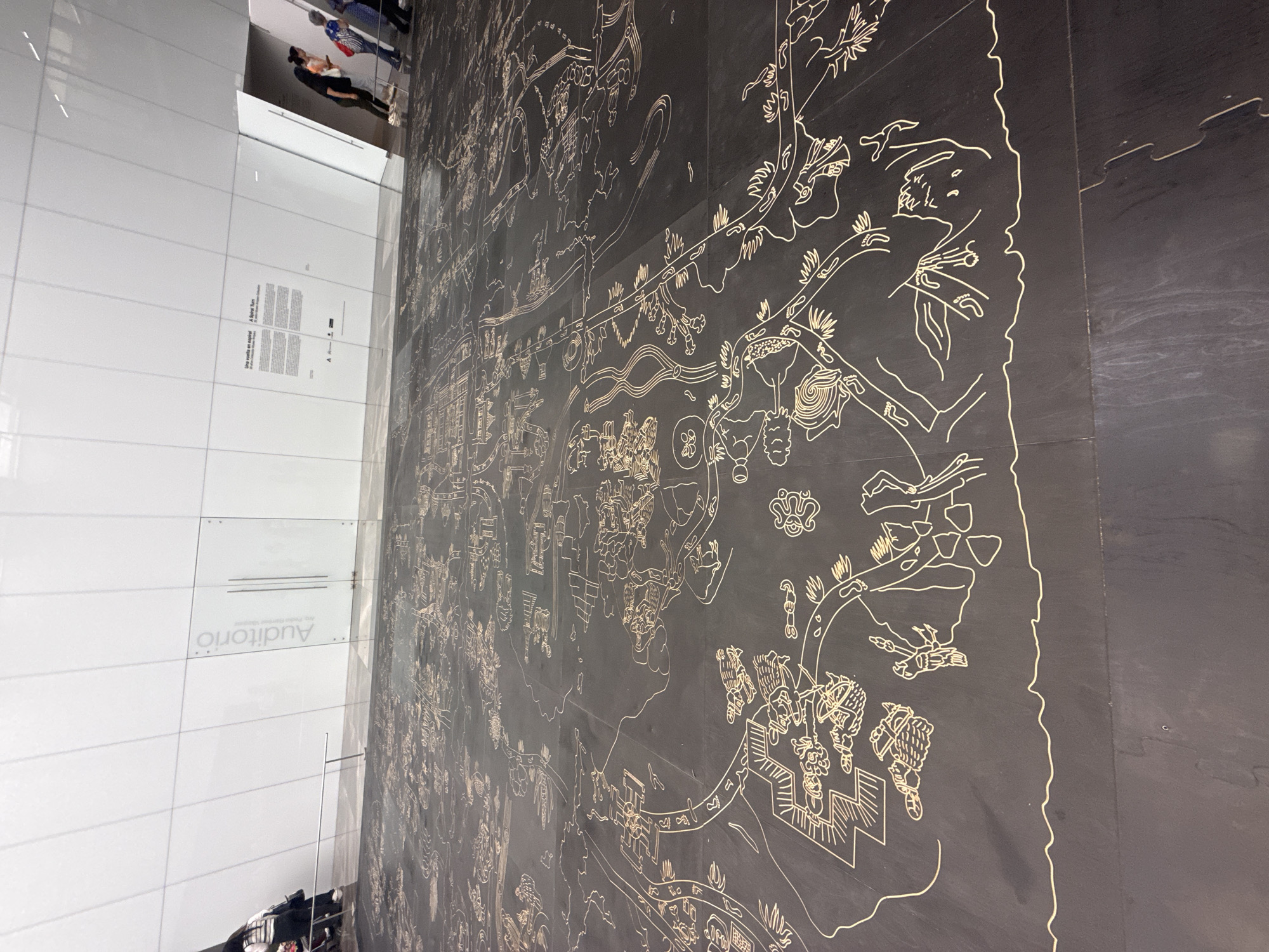 The image captures a scene within the 'Auditorio' of the International Museum of the Baroque in Puebla, Mexico. A large wall is adorned with a complex, light-colored linear drawing against a dark background. The drawing resembles a historical map or illustrative narrative, featuring elements like buildings, human figures, and natural motifs such as plants. To the left, a glass wall reflects the scene and the people outside the museum. The reflection shows people wearing casual clothes and walking near the museum. The reflective glass wall has the word 'Auditorio' written in a light colored font.
