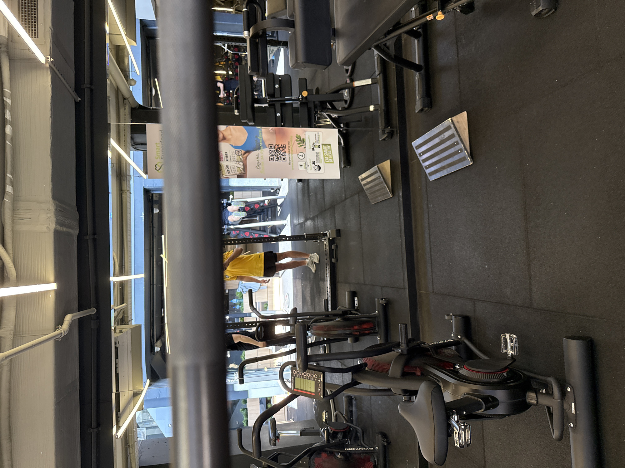 This indoor image shows a gym or fitness center interior. Various exercise machines are visible, including stationary bikes and weight lifting equipment. The focus is on the equipment and layout of the space. There is an individual wearing a yellow shirt and dark shorts walking in the background. The location and time of day are not discernable from the image. OCR text mentions the city of Sofia, Bulgaria.