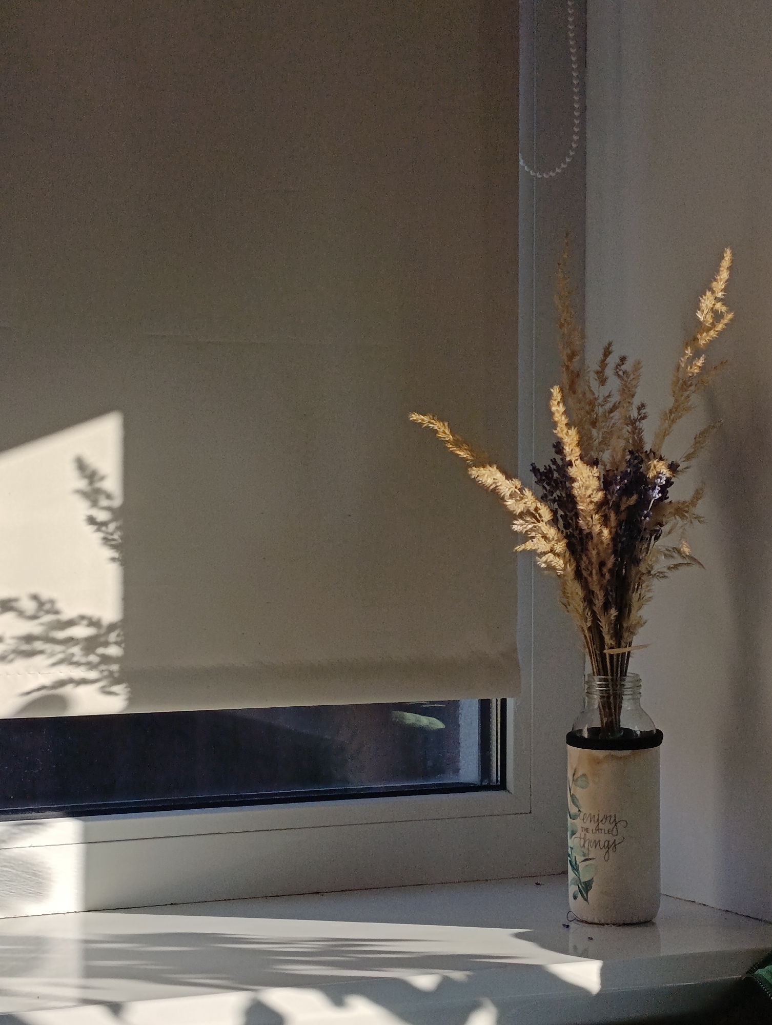 A sunlit windowsill holds a bouquet of dried grasses and lavender in a decorated glass bottle. The bottle has a textured, beige surface with green leafy designs and the words "enjoy the little things" in script. To the left, a window with a beige roller blind is partially open, revealing a dark interior. Sunlight casts a sharp shadow of branches on the blind and the wall to the left of the window. The scene is viewed from a slightly elevated angle, with the windowsill and the window frame in the foreground, and the wall and blind occupying the background.
