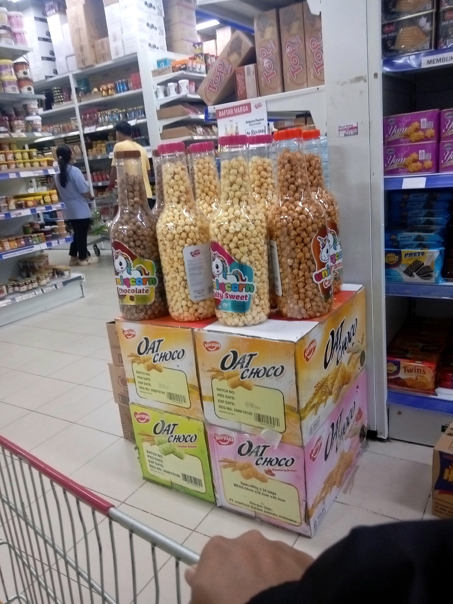 The image captures a view inside a retail store, possibly a supermarket or minimarket, in Batam, Indonesia. The foreground is dominated by the handle and part of the basket of a shopping cart, suggesting a shopper's perspective. Behind the cart, on a white tiled floor, are several stacks of boxed snacks. These boxes appear to be for "OAT CHOCO" brand, with labels indicating different flavors like "Pandan Pandan" and "Strawberry." Some of the boxes have a pink and white design, while others are yellow.
Above the snack boxes, on a tiered shelf, are three large plastic bottles filled with snacks. The leftmost bottle contains small, brown spherical items, and has a label with a cartoon unicorn and the text "Unicorn Chocolate." The middle bottle contains light brown, irregular shaped snacks, and has a similar unicorn label with text that appears to say "Unicorn Caramel." The rightmost bottle is filled with yellow, popcorn-like snacks and has a label with a cartoon character and "Salty Sweet."
Further in the background, shelves are stocked with various products. On the left, there are shelves with jars and cans of food items. On the right, a blue-lined shelf displays boxes of what appear to be cookies or biscuits, with labels including "PRESTO" and "YAM." Above these shelves, more boxes of products are stacked, with visible branding like "Top" and what looks like "DAFTAR HARGA" (price list) on a sign.
Two individuals are partially visible in the background. On the left, a person wearing a blue shirt and dark pants is walking away from the camera, pushing a shopping cart. On the right, a person wearing a yellow shirt and dark pants is also walking away. The overall environment suggests a well-stocked retail space with aisles of merchandise.