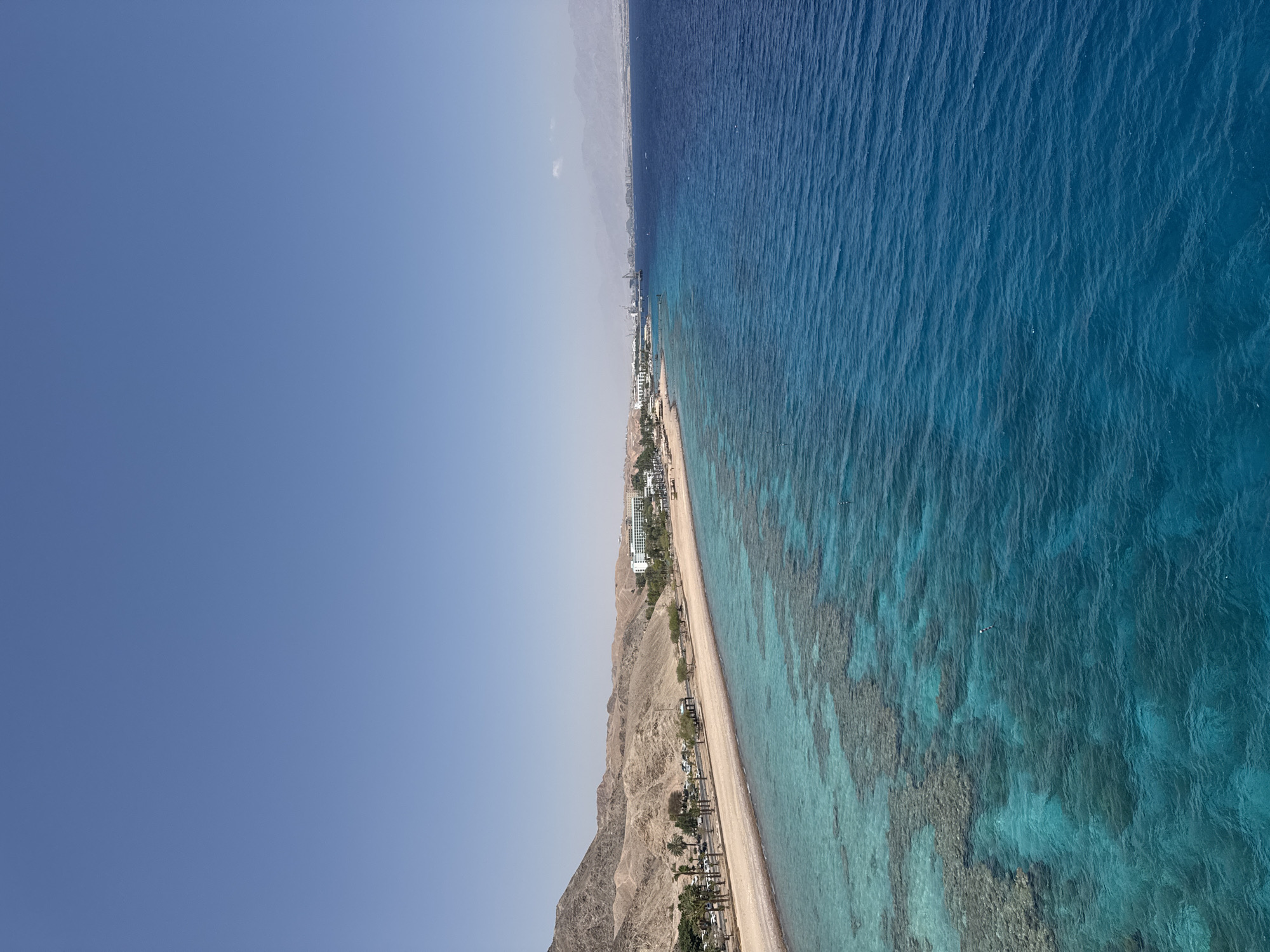 The image captures a panoramic view of a coastline in Eilat, Israel. In the foreground, clear turquoise waters reveal coral formations beneath the surface. The coastline features a sandy beach with a road running alongside it, lined with palm trees and parked cars. Beyond the beach, a range of desert mountains forms the backdrop. Several low-rise buildings, likely hotels or resorts, are visible along the shore, suggesting a developed tourist area. The vast expanse of the blue sky dominates the upper portion of the image. The sea is calm with a slight ripple, and the sky is clear, indicating a sunny day. The overall scene depicts a tranquil coastal resort environment with a blend of natural desert landscape and developed infrastructure.
