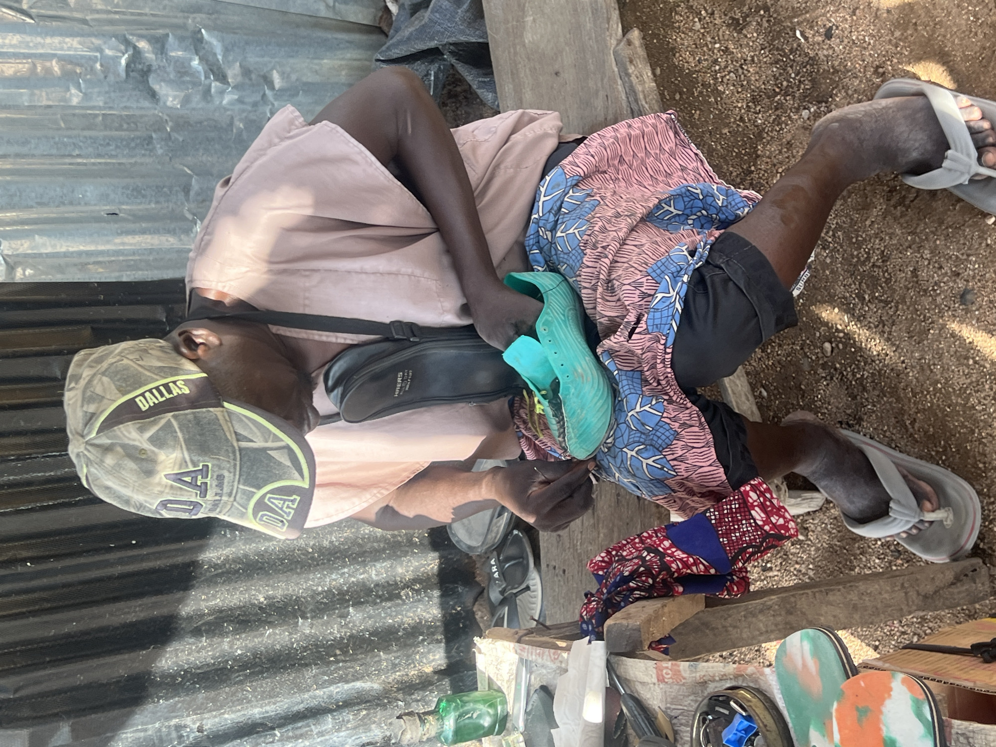 The image, taken from an overhead perspective, depicts a person engrossed in a task within an outdoor, informal setting. The individual, appearing to be an adult male with dark skin, is seated on a low wooden structure, framed by a background of corrugated metal sheeting and sandy ground.
He is dressed in a light pink/beige long-sleeved shirt, black shorts, and a colorful patterned fabric (blue, pink, and white motifs) draped around his lower body. A black shoulder bag, with "NHERS" and "ZARA" visible on it, is worn across his chest. On his head, he wears a camouflage-patterned baseball cap displaying "DALLAS" and "DA" in lime green and purple. His right foot is in a grey flip-flop, which also shows the word "ZARA" on its strap. The person's attention is fixed on a bright turquoise/light green shoe held in his left hand, suggesting he is working on it, possibly repairing or inspecting it.
The scene is brightly lit by natural sunlight, indicating daytime. Surrounding objects include wooden planks, a dark tarp, and various items on a surface beside him. These items consist of a green glass bottle, colorful textiles featuring red, blue, and white patterns, and several sandals with distinctively patterned soles in green, orange, and white. The setting, with its corrugated metal walls and sandy ground, suggests a street market or a similar commercial outdoor space.