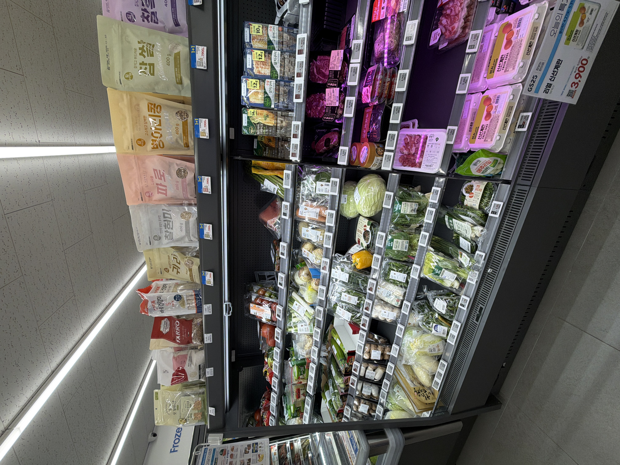 The image depicts the interior of a supermarket in Seoul, South Korea. The focus is on refrigerated shelves stocked with various food items.
On the left side, there are packages of grains and beans, including rice, barley, and beans. Brands like "FARRO" and products labeled "Glutinous Rice" are visible.
The central shelves hold produce, including packaged vegetables like cabbages, scallions, lettuce and mushrooms. Yellow bell pepper is also visible.
To the right of the produce, the shelves contain packaged meat products and eggs. Boxes of eggs labeled "Fresh Eggs" are prominently displayed.