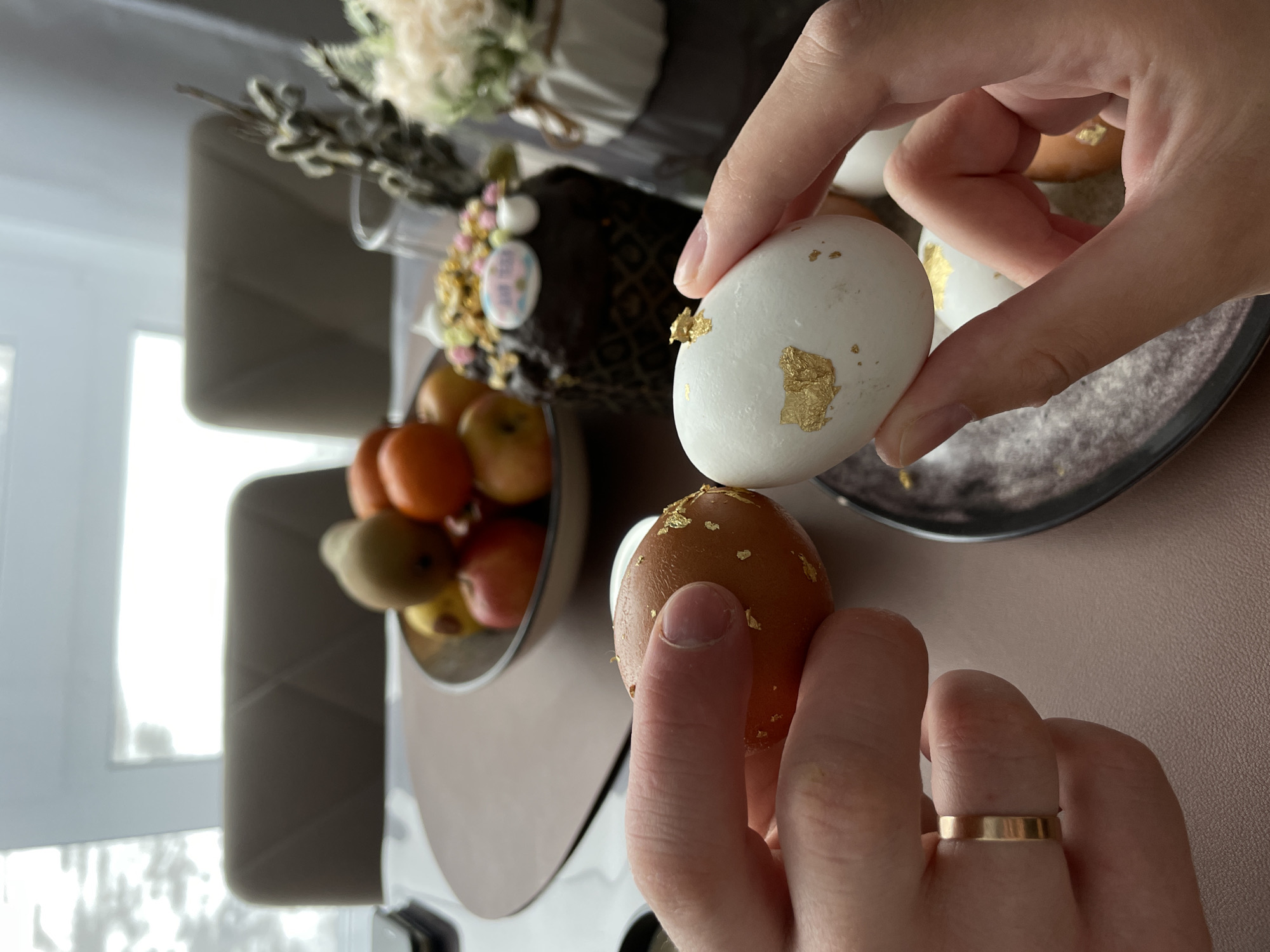 The image depicts a close-up shot of two hands holding a brown egg and a white egg. Both eggs are decorated with small pieces of gold leaf. The hand holding the brown egg has a gold ring on one finger. Behind the hands and eggs is a blurred background of a table with a bowl of fruit, a decorated cake, and possibly some flowers. The scene appears to be indoors. Based on the prompt, the location is Подшивалово, Russia.