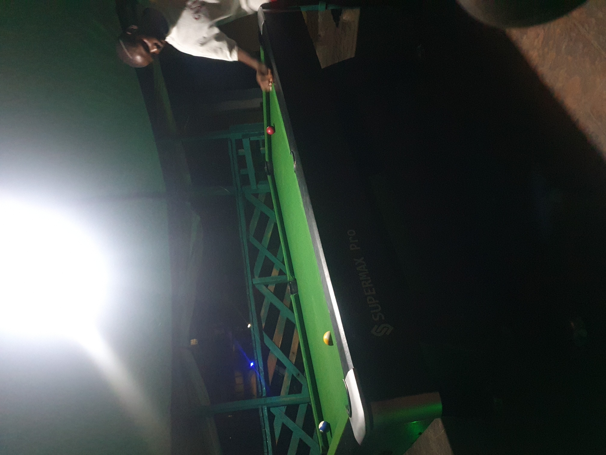 The image depicts an outdoor recreational area in Lokoja, Nigeria, at night. A pool table is the central focus, illuminated by overhead lighting that creates a bright glare. A Black male, wearing a white t-shirt, stands to the right of the pool table, his hand extended towards it, suggesting he is playing. The pool table itself is green with various colored balls, including a red ball, a yellow ball, and a striped ball. A blue light is visible in the background, emanating from what appears to be a railing or fence structure. The environment is dark, with the pool table and the surrounding structures providing the only significant illumination. The overall impression is of a casual, nighttime gathering or activity.
