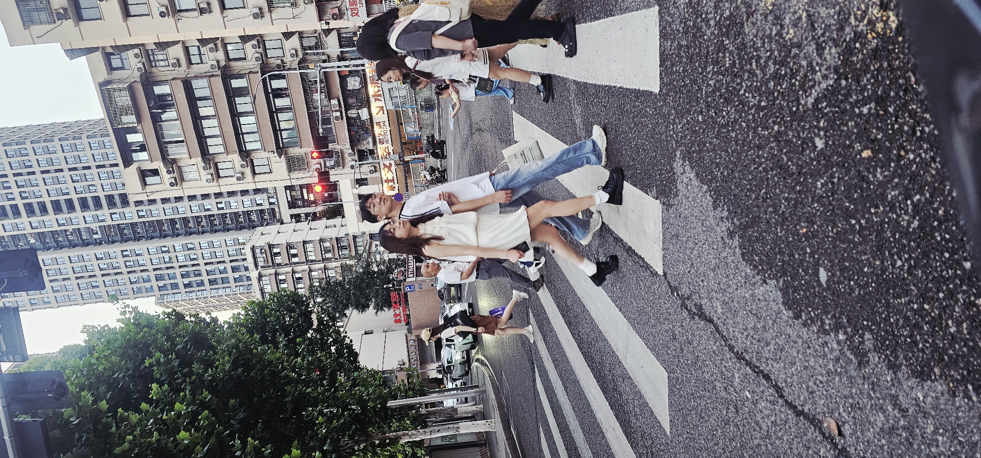 The image depicts a street scene in Yuzhong District, China. Several pedestrians are crossing a zebra crossing. Among them are a woman in a white dress and a man in a white t-shirt and jeans, walking closely together. Other pedestrians are nearby, including a group of people to their right and others further back. The buildings are typical of an urban environment with multiple stories and rows of windows. The street surface is asphalt, and there are traffic lights visible. The lower left corner includes green foliage, likely from trees lining the street. Cars are parked along the roadside, and the overall lighting suggests either early morning or late afternoon.