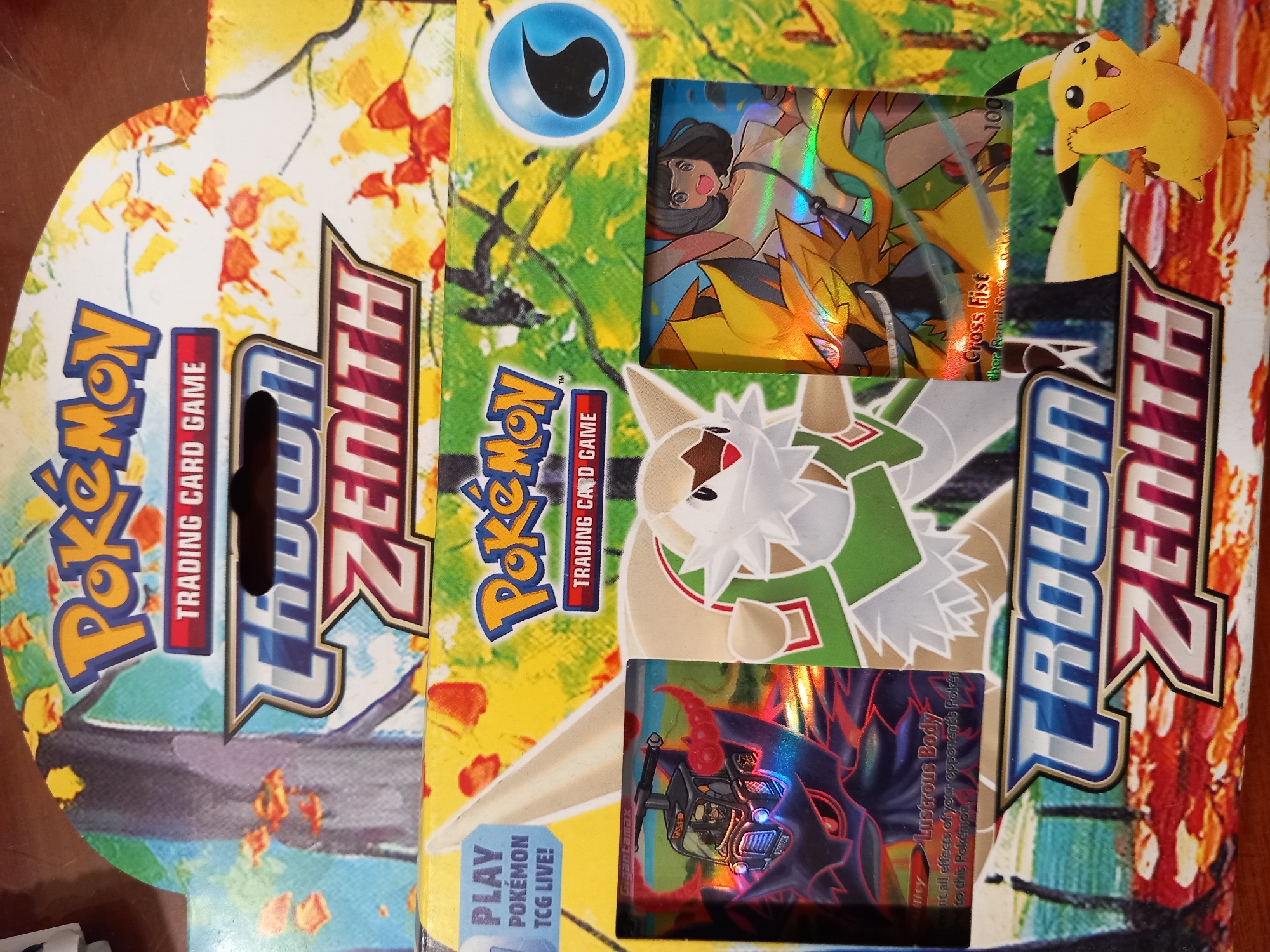 The image displays a collection of Pokémon trading cards and a box, all presented in a top-down, slightly angled view. The primary subjects are the colorful Pokémon cards and the "Crown Zenith" themed box, which features vibrant artwork with autumn-colored foliage and imagery of Pokémon characters.
The cards themselves are the focal point. One card prominently features a character with black hair and large eyes, wearing a white top, with a yellow and orange Pokémon in the background. Another card shows a Pokémon with white and brown fur, green accents, and red eyes. The third visible card showcases a dark-colored Pokémon with red horns and a metallic, glowing quality. Text on the cards includes "Cross Fist," "Gigantamax," and "Lustrous Body," as well as what appears to be a health point value of "100."
The "Crown Zenith" box has the Pokémon logo and "Trading Card Game" printed on it, along with the "Crown Zenith" branding in large, stylized letters. A small, cheerful Pikachu figure is positioned on the top right corner of the box. The overall scene appears to be an organized display of these Pokémon-related items, possibly for sale or as part of a collection. The lighting is bright, suggesting it was taken in a well-lit indoor environment, though no specific location cues are present beyond the items themselves. The colors are rich and saturated, highlighting the playful and energetic nature of the Pokémon brand.