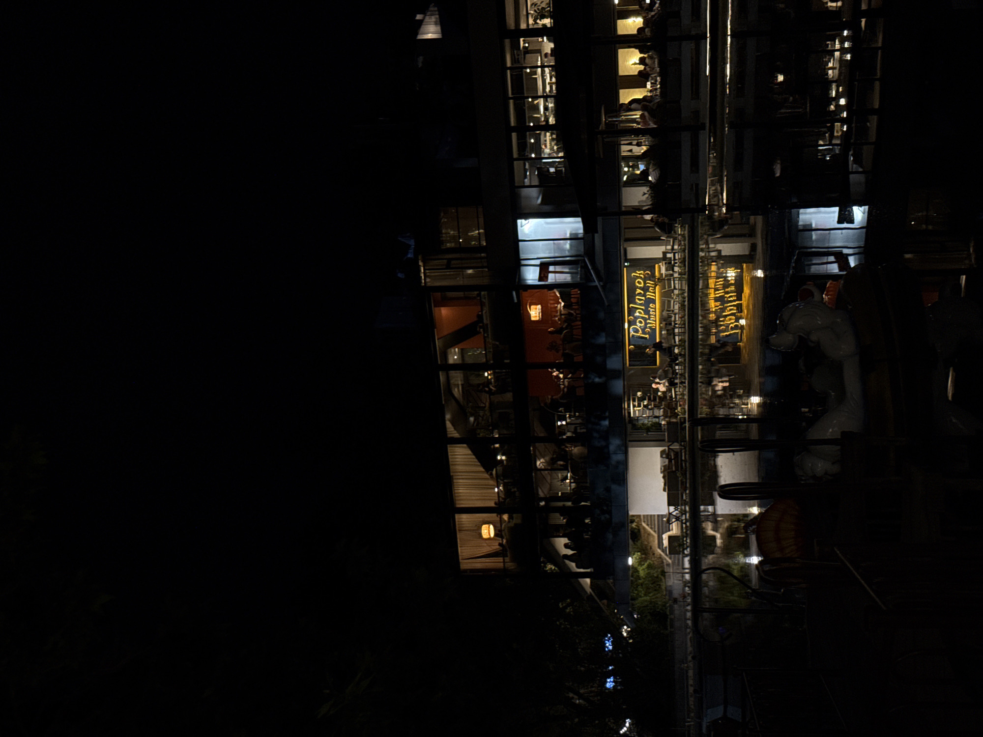 The image displays a multi-story building at night, brightly illuminated against the dark urban backdrop. The structure features numerous large glass windows, revealing various indoor spaces. Inside, indistinct figures of people can be seen seated at tables, suggesting activities such as dining, drinking, or socializing within multiple establishments.
A prominent yellow, illuminated sign on a lower level of the building clearly displays the text "Poplavok Music Hall," accompanied by a stylized graphic. Below this sign, several large, white, artistic sculptures or figures are visible on the exterior ground level. Another distinct, large orange or yellow lantern-like fixture hangs on the building's facade. The foreground is dark, showing subtle outlines of foliage or trees.
The scene depicts a lively evening environment, with the building's interior lights providing the primary source of illumination. The time of day is clearly night. The visible text "Poplavok Music Hall" serves as a specific location cue, situating the scene in Yerevan, Armenia, indicating a place of entertainment or dining. The overall atmosphere suggests leisure and social activity.