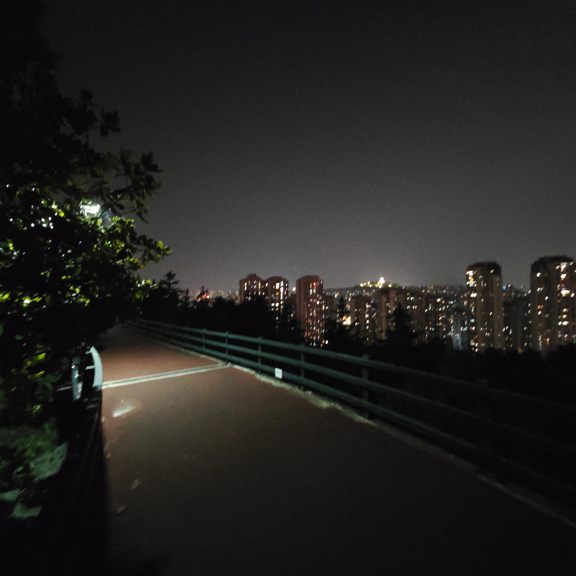 The image shows an elevated walkway with a red surface, likely a running track, at night in Yuzhong District, China. A dark green railing runs along the right side of the walkway. The walkway is poorly illuminated, transitioning into deep shadow in the foreground. A tree, brightly lit by a lamp, occupies the left side of the image. In the background, a cityscape of high-rise buildings with numerous illuminated windows is visible, suggesting a densely populated urban area. The sky is overcast and dark.