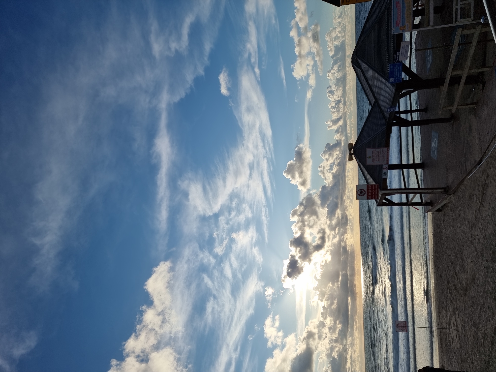 The image captures a late afternoon or early evening scene on a beach in Tel-Aviv, Israel, with the sun positioned low over the western horizon, casting a bright glare across the Mediterranean Sea. The sky is predominantly blue with numerous white and grey cloud formations, including both fluffy cumulus and wispy cirrus clouds. Sunbeams radiate through gaps in the clouds above the horizon.
The foreground consists of sandy beach leading to the ocean, which displays moderate waves with visible white crests. On a raised wooden platform near the shoreline, two dark-roofed, open-sided wooden shelters, possibly gazebos, are present. The left shelter features a prominent red-bordered warning sign with Hebrew text and a "no diving" pictogram. Two horn-shaped speakers are mounted on the peak of this shelter's roof. Another red-bordered sign with Hebrew text is visible on a pole planted in the sand to the left of this shelter. The right shelter also has multiple signs affixed to its wooden posts, displaying Hebrew text and accessibility symbols, including a wheelchair icon. A wooden railing runs along the right side of the platform.
A single individual, clad in dark attire, is partially visible on the far left edge of the frame, standing on the sand and facing the sea, mostly obscured by the left shelter's structure.