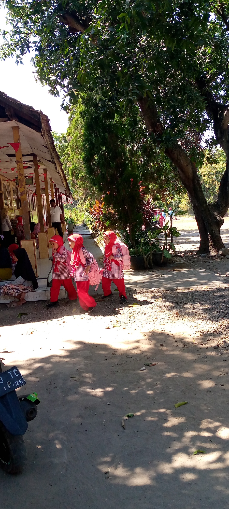 In Pacing, Indonesia, three young girls in red hijabs and checkered uniforms with pink backpacks walk down a dirt path lined with trees. To their left, a building with a red-and-white striped awning has several people sitting and standing near its entrance. A motorcycle is parked in the foreground, on the left side of the frame. The scene is brightly lit by the sun, casting shadows from the trees onto the ground.