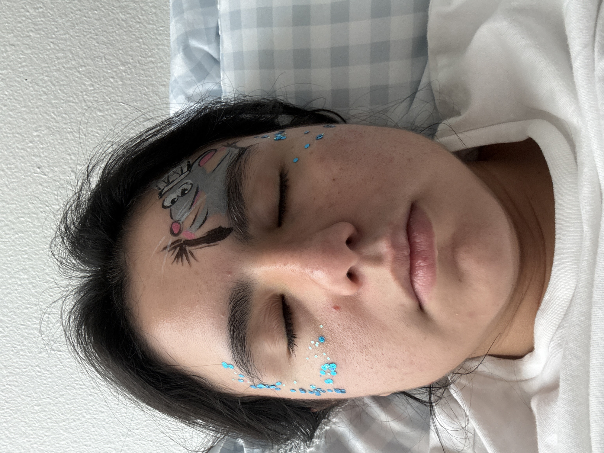 A close-up, overhead shot shows a person lying in bed with their eyes closed. The person has dark hair and is wearing a white t-shirt. There is glitter applied to the left side of their face, and face paint of a cartoon character on their forehead. The background consists of a white textured wall and a blue and white checkered pillow. The image was taken in Austin, United States of America.