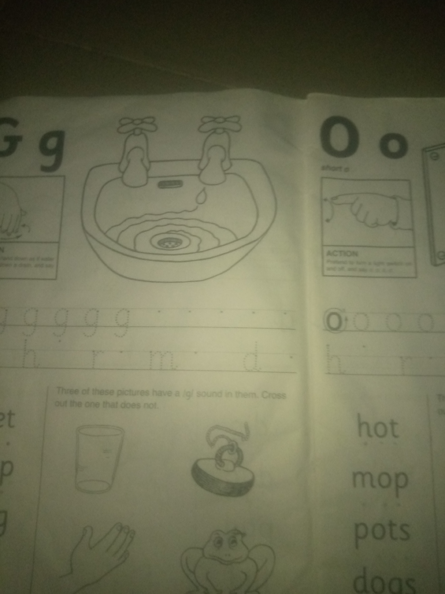 The image shows a page from a children's workbook, likely for learning the alphabet and phonics. The page is divided into sections. On the left, the letters "G g" are displayed, followed by a drawing of a sink with two faucets, and some dotted letters "g". Below this are dotted letters "h", "r", and "m". A section asks the reader to identify three pictures with a /g/ sound and cross out the one that does not. Three pictures are shown: a glass, a toy that looks like a bell or spinning top, and a hand.
On the right side of the page, the letters "O o" are displayed with the text "short o", followed by a drawing illustrating an action and the word "ACTION". Below this are dotted letters "O" and other circled letters. Further down are words written in a larger font: "hot", "mop", "pots", and "dogs".
The image is taken from a perspective that suggests the workbook is lying on a surface, possibly a table or desk, with a dark, textured background, possibly a floor or wall. The lighting is uneven, with some areas brighter than others, suggesting an indoor setting with artificial light. The overall impression is of a classroom or home learning environment. The image does not contain any specific geographical markers that would indicate its location in Jauro Yinu, Nigeria.