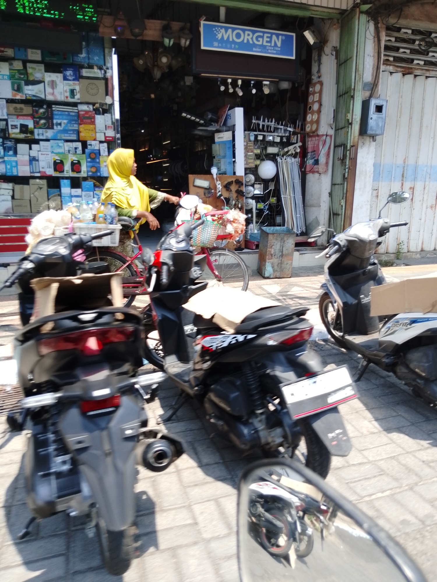 A street scene in Pasuruan, Indonesia, shows a woman wearing a yellow hijab standing next to a bicycle laden with goods. She is partially obscured by several parked motorbikes. The motorbikes are dark colored and modern in appearance. Behind the woman is a shop with an open entrance displaying various items, including light fixtures and boxes of products. A blue sign with the word "MORGEN" is visible above the shop entrance. The ground is paved with grey bricks, and the lighting suggests it is daytime with bright sunlight casting shadows. The overall impression is a bustling commercial street.