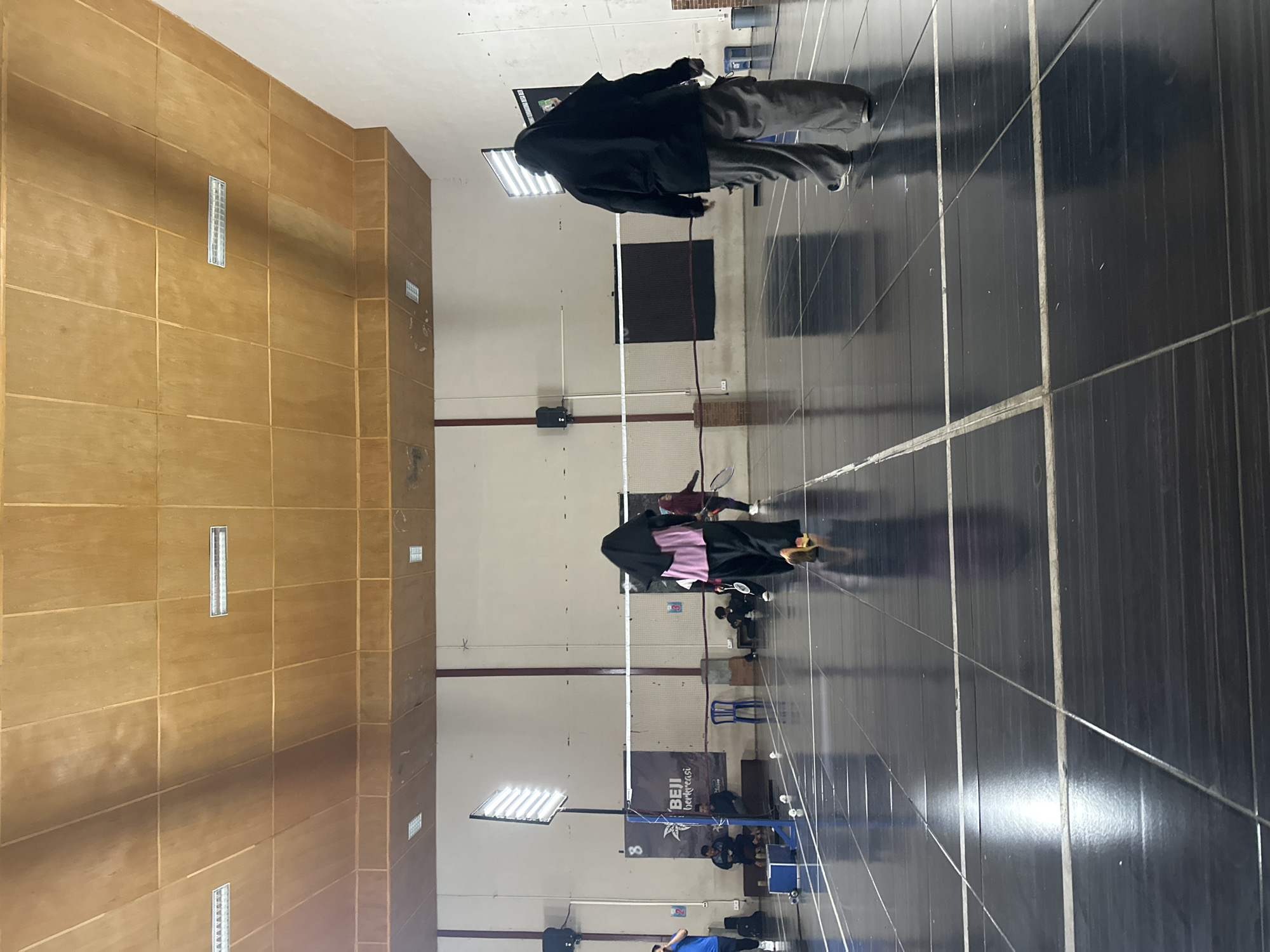 This image depicts an indoor badminton court, likely within a facility in the city of Batu, Indonesia. The court has a dark, reflective tiled floor with white lines demarcating the playing area. A white net stretches across the middle of the court. Several individuals are present. In the foreground, from behind, a person wearing a black hijab and a long black top with grey baggy pants walks away from the camera towards the net. Another person wearing a pink hijab and a long black garment is also moving away from the camera. To the right, another individual in a black hijab and grey pants is visible in mid-court, also facing away. Further back, near the left side of the court, several individuals are seated, and one person in a blue shirt appears to be standing. A large, bright light fixture on a stand is positioned on the left side of the court. The ceiling is made of wooden panels, and fluorescent lights are embedded in it. A banner with text and a logo is visible on the left side of the court, behind the net. The overall impression is a casual badminton game or practice session taking place in a well-lit indoor sports venue.