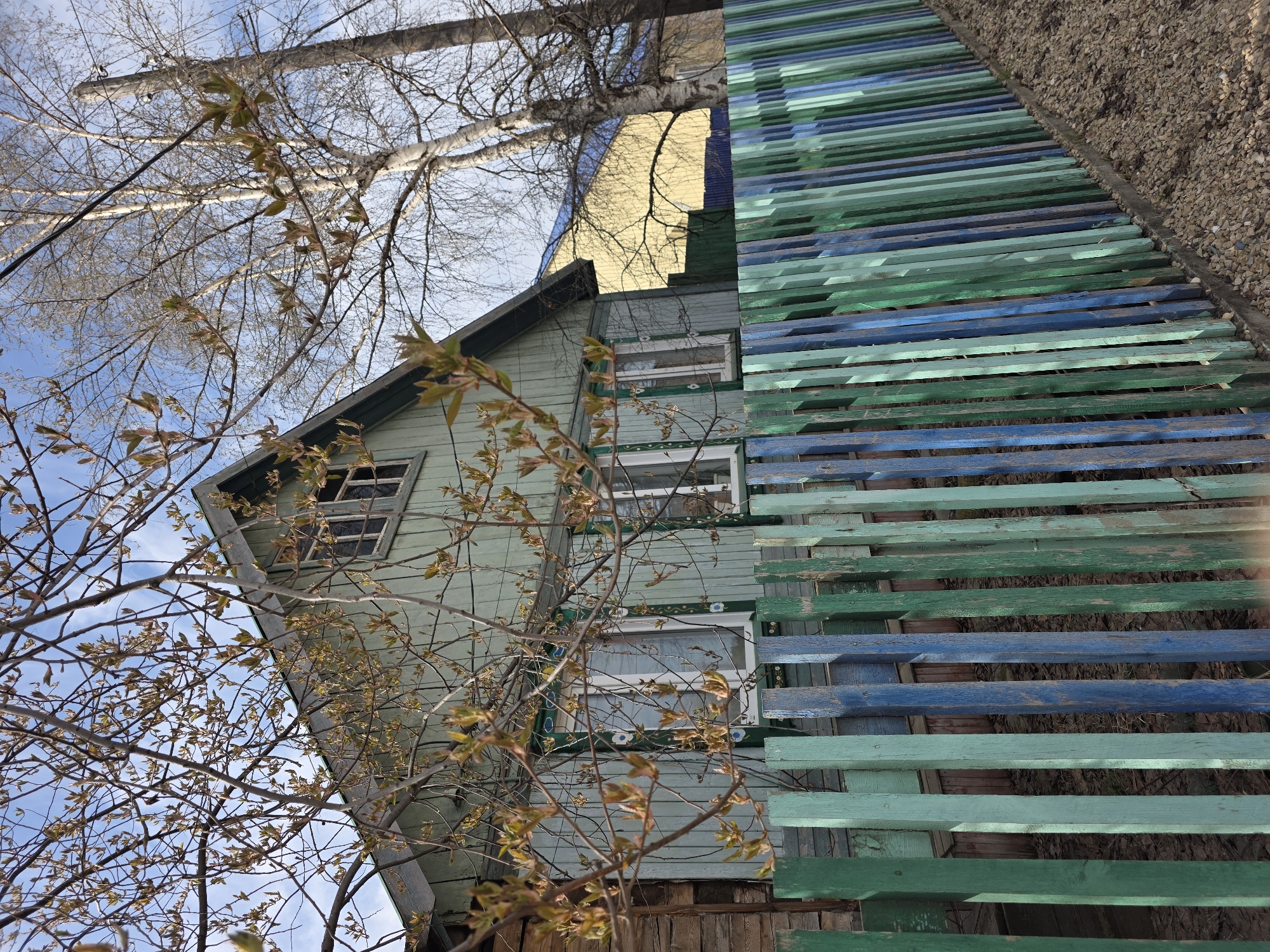 The image shows a two-story light-green wooden house in Lensk, Russia. The house has white-framed windows, some with decorative detailing. The upper story has a visible gable roof. The house is partially obscured by bare tree branches with budding leaves. Adjacent to the house, on the right side of the image, is a slatted structure made of painted wooden planks in alternating shades of green and blue. The structure appears to be a fence or retaining wall, with gravel visible behind it. The sky is visible in the background, showing blue patches interspersed with light clouds. A tree branch stretches across the top of the image.