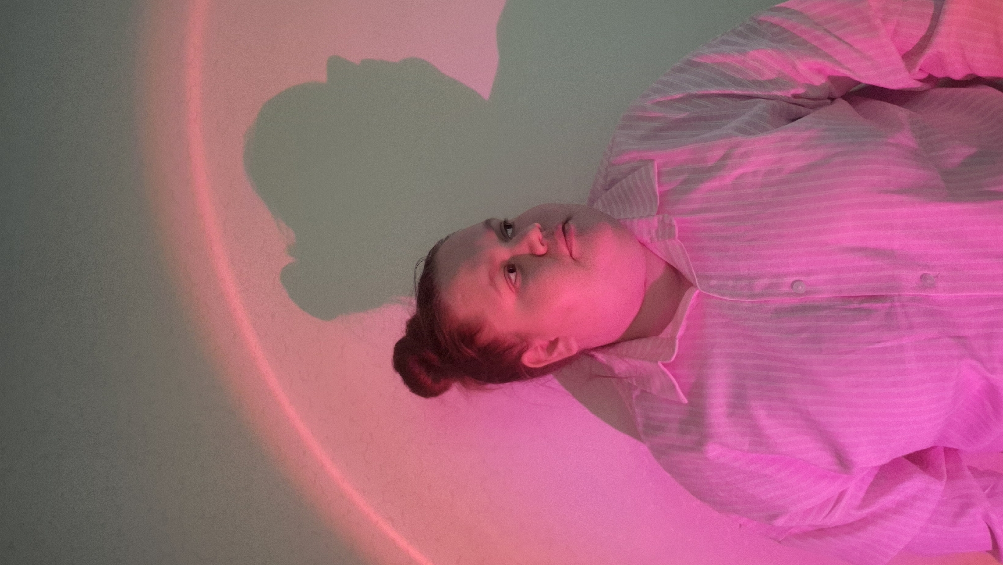 A woman in a pink and white striped shirt lies on her side, bathed in pink light. Her hair is pulled back into a bun. A shadow, seemingly of her head and shoulders, is cast on the wall beside her, intersecting with a pink light band. The environment suggests an indoor setting. No specific details indicate the location as Chaikovsky, Russia.