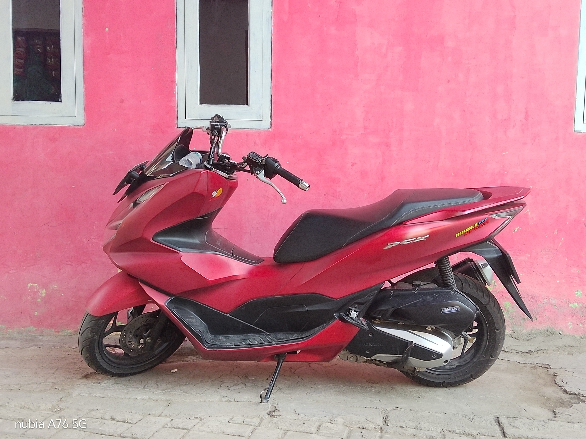 A red Honda PCX scooter is parked on a paved surface in front of a bright pink wall. The wall has two white-framed windows, one on the left and one on the right, with dark panes. The scooter is viewed from its left side, with its front wheel on the left and its rear wheel on the right. The scooter has a black seat, black footrest area, and a black exhaust. A small kickstand is deployed, holding the scooter upright. The ground is made of interlocking brick pavers. In the left window, a reflection shows a person wearing a green headscarf and shelves with items. A watermark in the bottom left corner of the image reads "nubia A76 5G". The overall scene is outdoors, possibly in a residential or commercial area in Cakung, Indonesia, given the presence of the windows and the scooter.