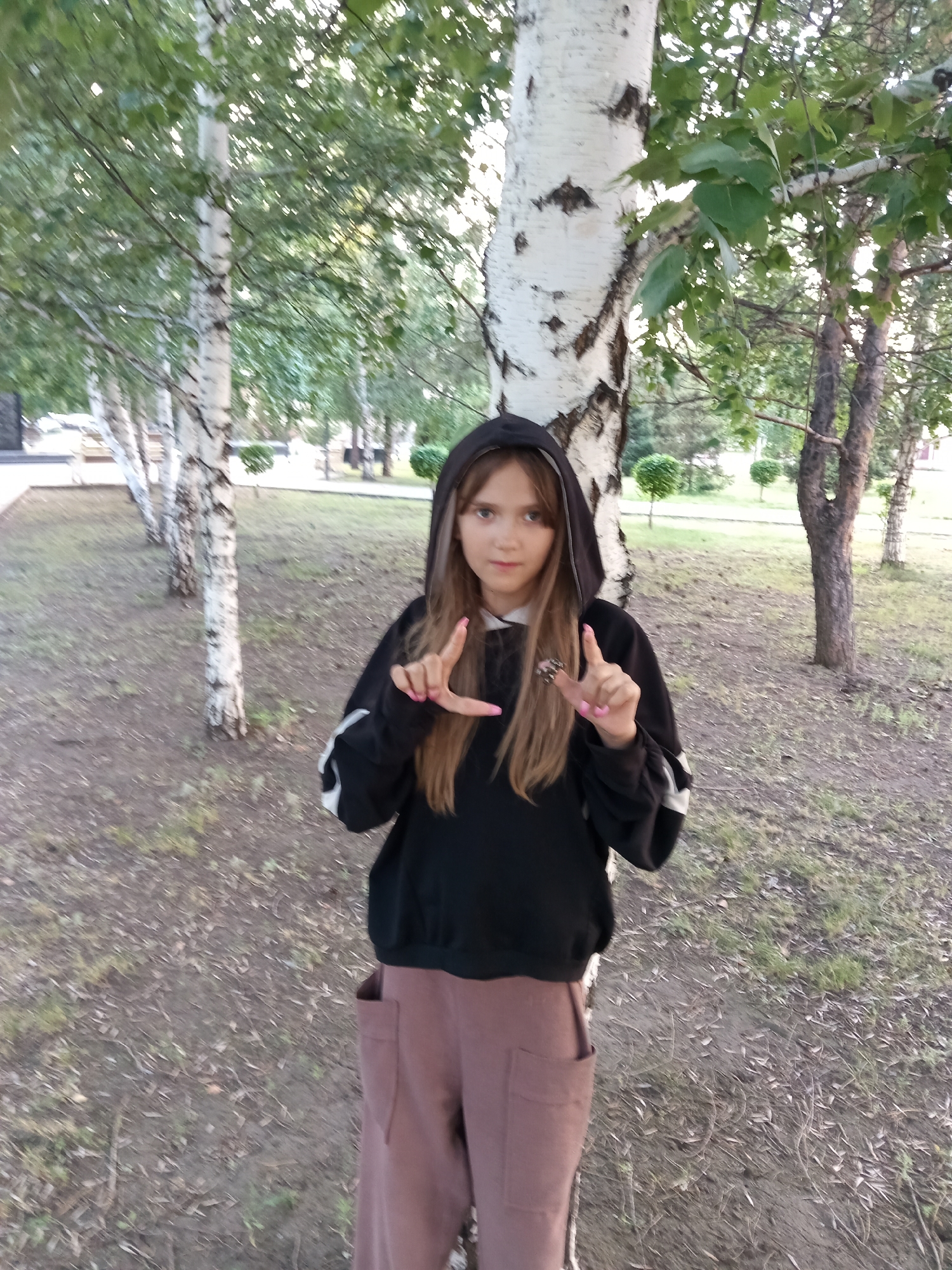 In Rudny, Kazakhstan, a young girl with long, light brown hair and light brown eyes stands in a park-like setting. She is wearing a black hoodie with white stripes on the sleeves and loose, brown pants. Her nails are painted pink. She is making a "L" shape with her left hand and a peace sign with her right hand. The background features a row of birch trees with white bark and green leaves, and a dirt path with fallen leaves. The sun is casting dappled light through the trees.