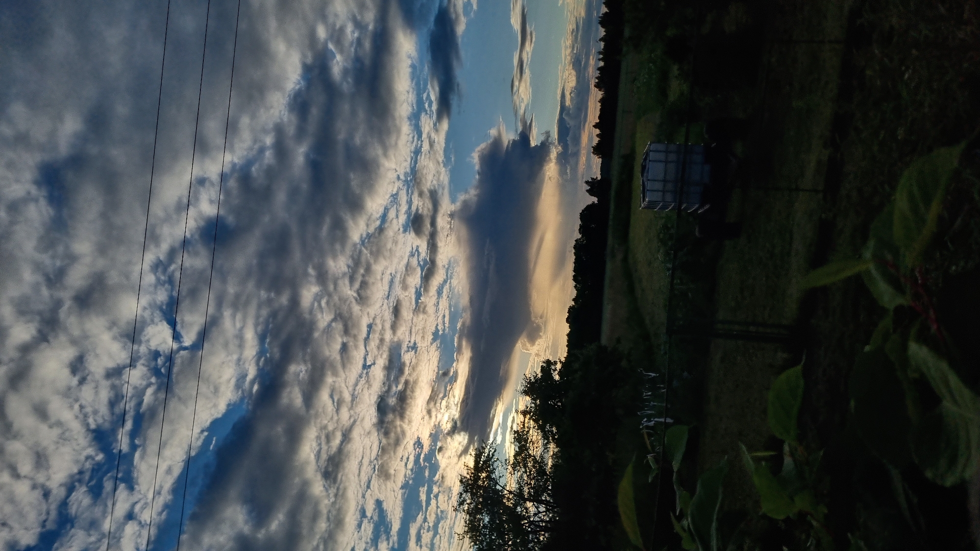 The image depicts a twilight rural landscape in Vysokoe, Russia, under a dramatic sky filled with dark, textured clouds interspersed with patches of bright blue and the setting sun's warm glow. In the foreground, dark green foliage occupies the lower left corner, with thin red stems visible. Behind this, a clothesline with several items hanging on it stretches across the middle ground. Beyond the clothesline, a dark green field is visible, extending towards a line of silhouetted trees on the horizon. A white cube-shaped agricultural container is positioned in the field, resting on what appears to be a trailer or vehicle. Three dark, horizontal power lines traverse the upper portion of the frame against the cloudy sky. The overall impression is one of a serene, yet dynamic, natural scene at dusk.