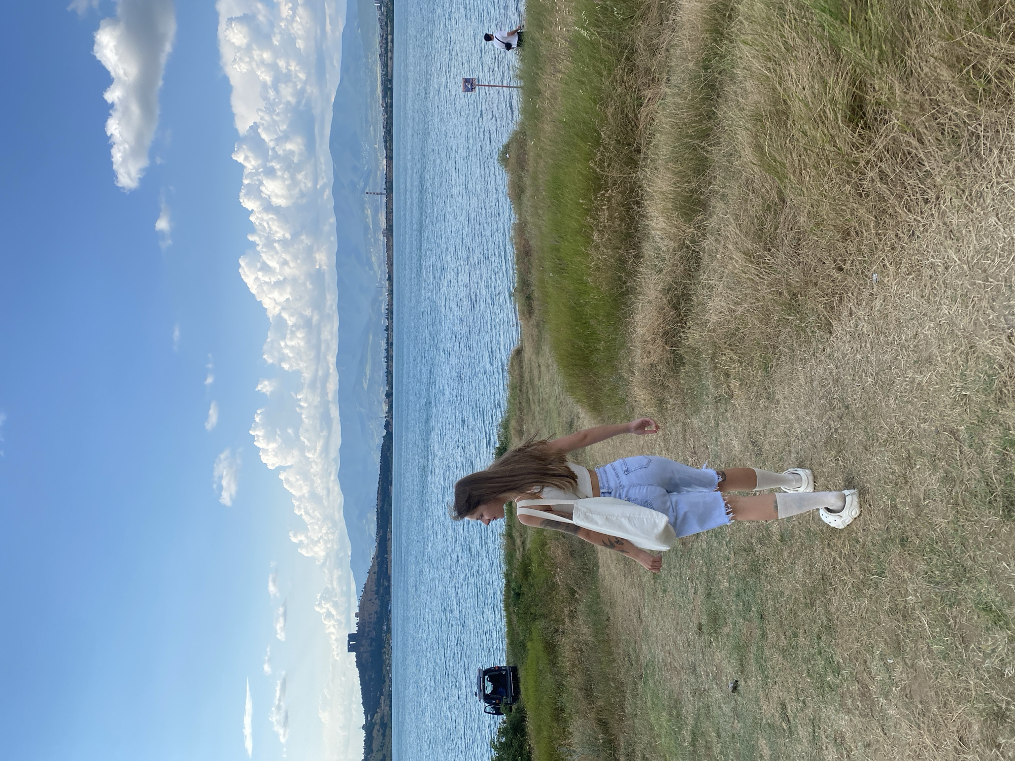The image depicts a woman from behind, walking on a grassy path towards a large body of water. She is wearing a white crop top, light blue denim shorts, white socks, and white clogs. A white tote bag hangs from her shoulder. In the background, there is a vast blue body of water with ripples, and across the water, a hilly landscape is visible with a prominent fortress-like structure on top of a hill. The sky is a bright blue with scattered white clouds. To the left, a dark-colored vehicle with its rear door open is parked near the water's edge. Further to the right, along the shore, a man wearing a white t-shirt and dark shorts is visible, standing near a red signpost. The overall environment appears to be a scenic outdoor location, possibly near a reservoir or lake, with hills and a distant urban area. The image does not provide specific details to pinpoint the location within Tbilisi, Georgia.