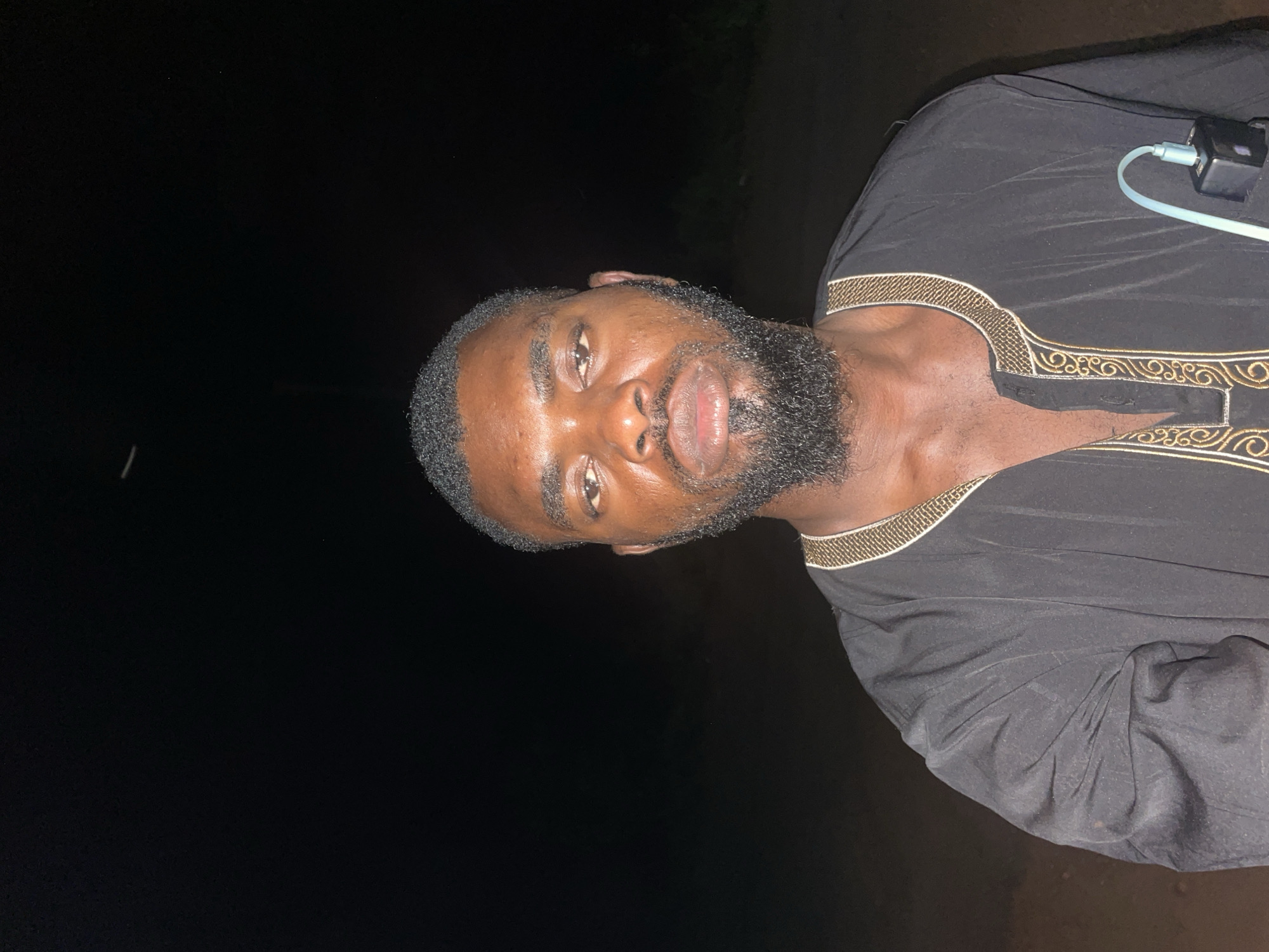 A man with a beard and dark hair is depicted in a photo taken at night in Ogbe, Nigeria. He's wearing a black shirt with gold trim around the collar and a charging block is attached to his shirt. The background is dark, with indistinct foliage visible above the man.