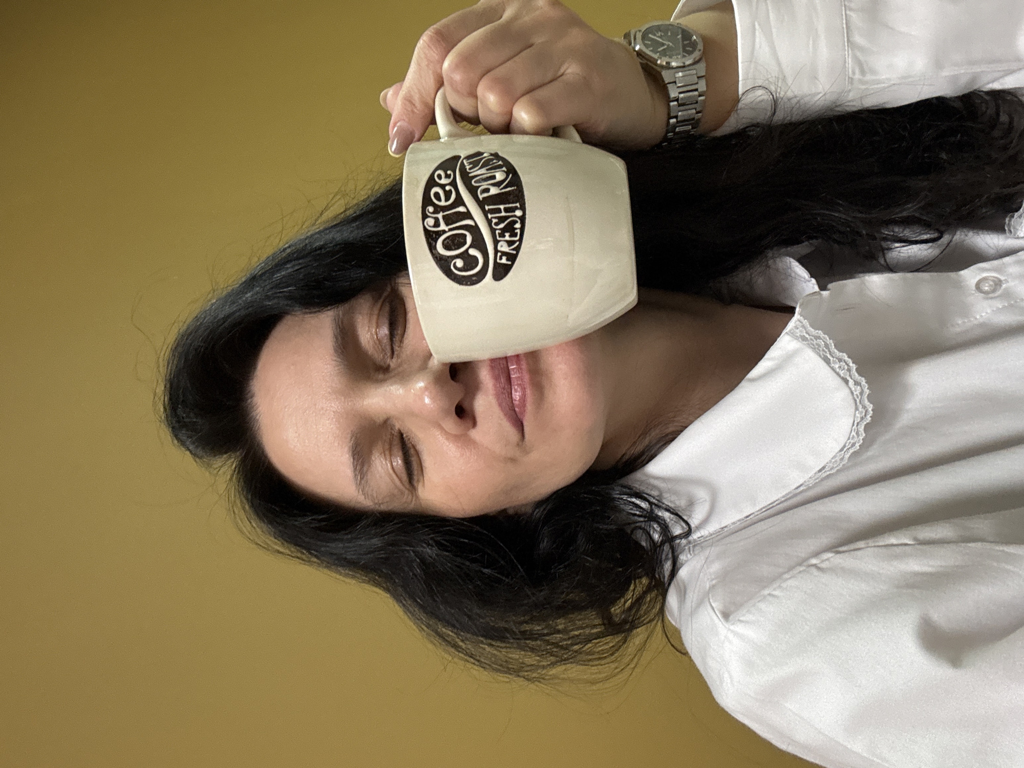 A woman is holding a white mug with the words "Coffee Fresh Roast" printed on it to her eye. The woman has long dark hair and is wearing a white shirt and a silver watch. The background is a solid yellow color. There is no information to confirm the location is Chaikovsky, Russia.