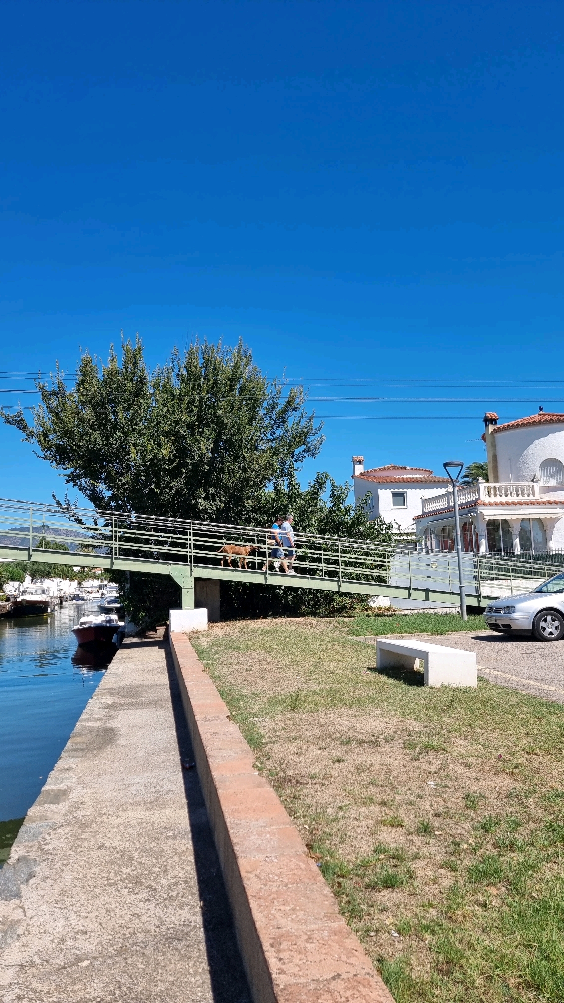 The image depicts a clear blue sky over a canal in Castelló d'Empúries, Spain. A pedestrian bridge spans the canal, painted green and supported by concrete pillars. A person wearing a blue t-shirt and shorts is walking across the bridge, accompanied by a medium-sized brown dog. The bridge has a metal railing. On the right side of the canal, there is a grassy area with a white concrete bench. Beyond the grass, a silver car is parked, and white houses with red-tiled roofs are visible. Boats are docked in the canal to the left of the bridge. The canal bank is a concrete walkway bordered by a low brick wall.