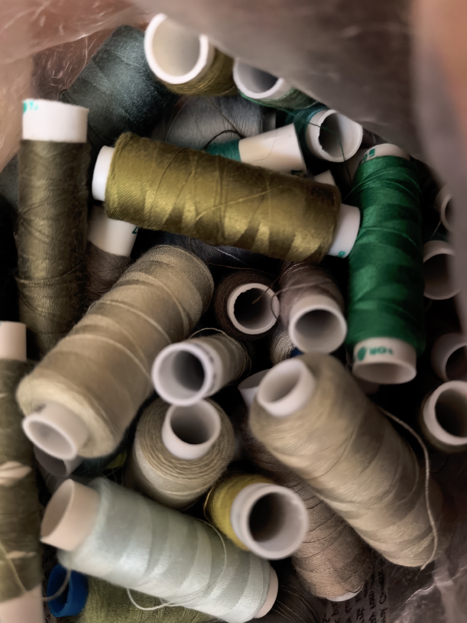 This is a close-up, overhead shot of a collection of spools of thread inside a plastic bag. The threads are in various shades of green, beige, and brown, with some spools appearing lighter and others darker depending on the color and lighting. The spools are cylindrical with white or light-colored plastic caps at each end. The threads are tightly wound around the spools. The background is blurry and appears to be the inside of the plastic bag, with some texture visible. There is no indication of the city of Sterlitamak, Russia, or any people or actions within the image.