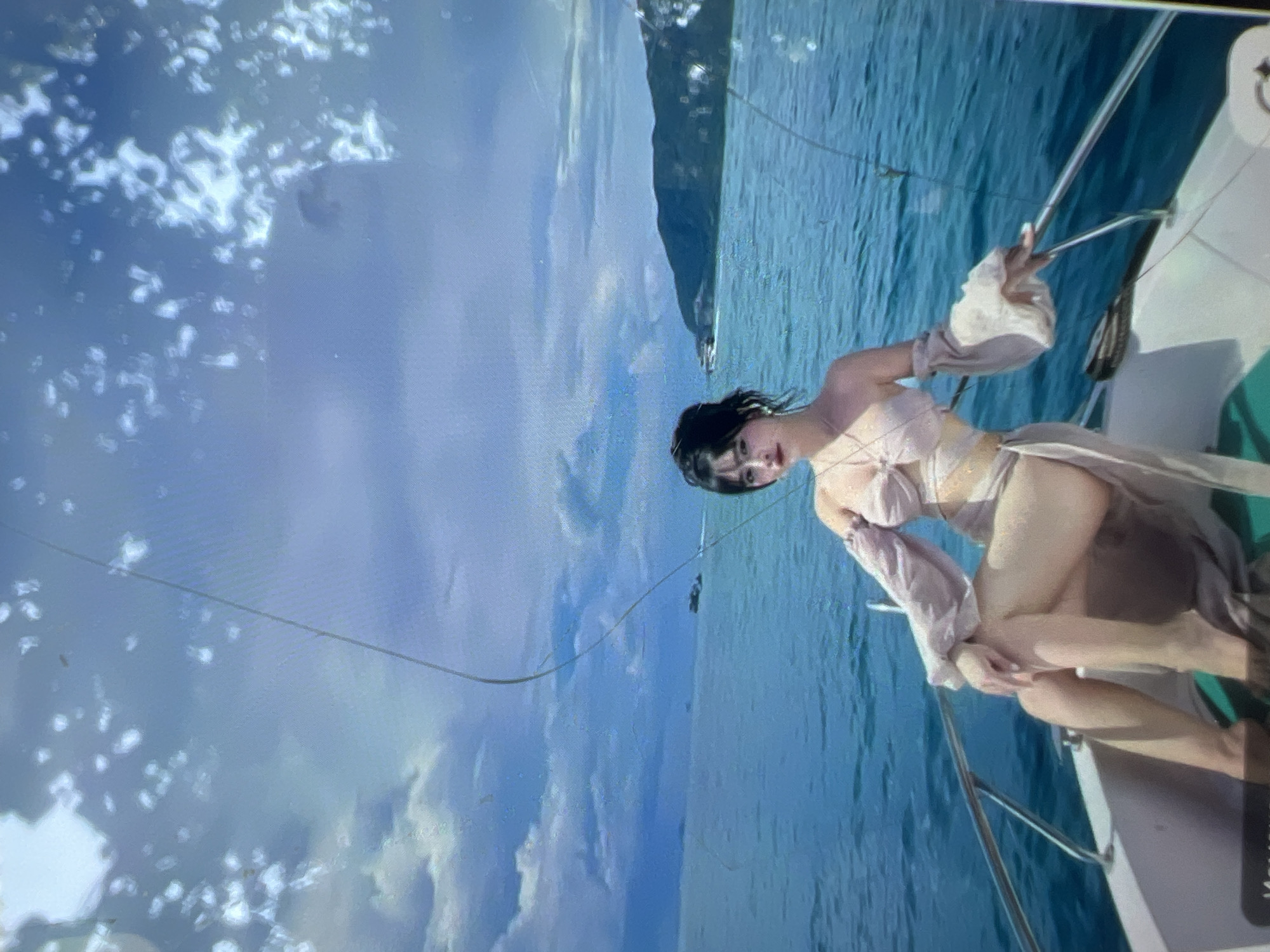 A young woman with dark hair and red lipstick sits on the bow of a white boat, her legs crossed. She wears a light-colored bikini top and a sheer skirt that drapes around her. Her arms are adorned with voluminous, bell-shaped sleeves. The boat sails on a turquoise sea under a cloudy sky. In the distance, a green, mountainous coastline is visible. There are no elements in the image that indicate a location in Kara-Dıykan, Kyrgyzstan.