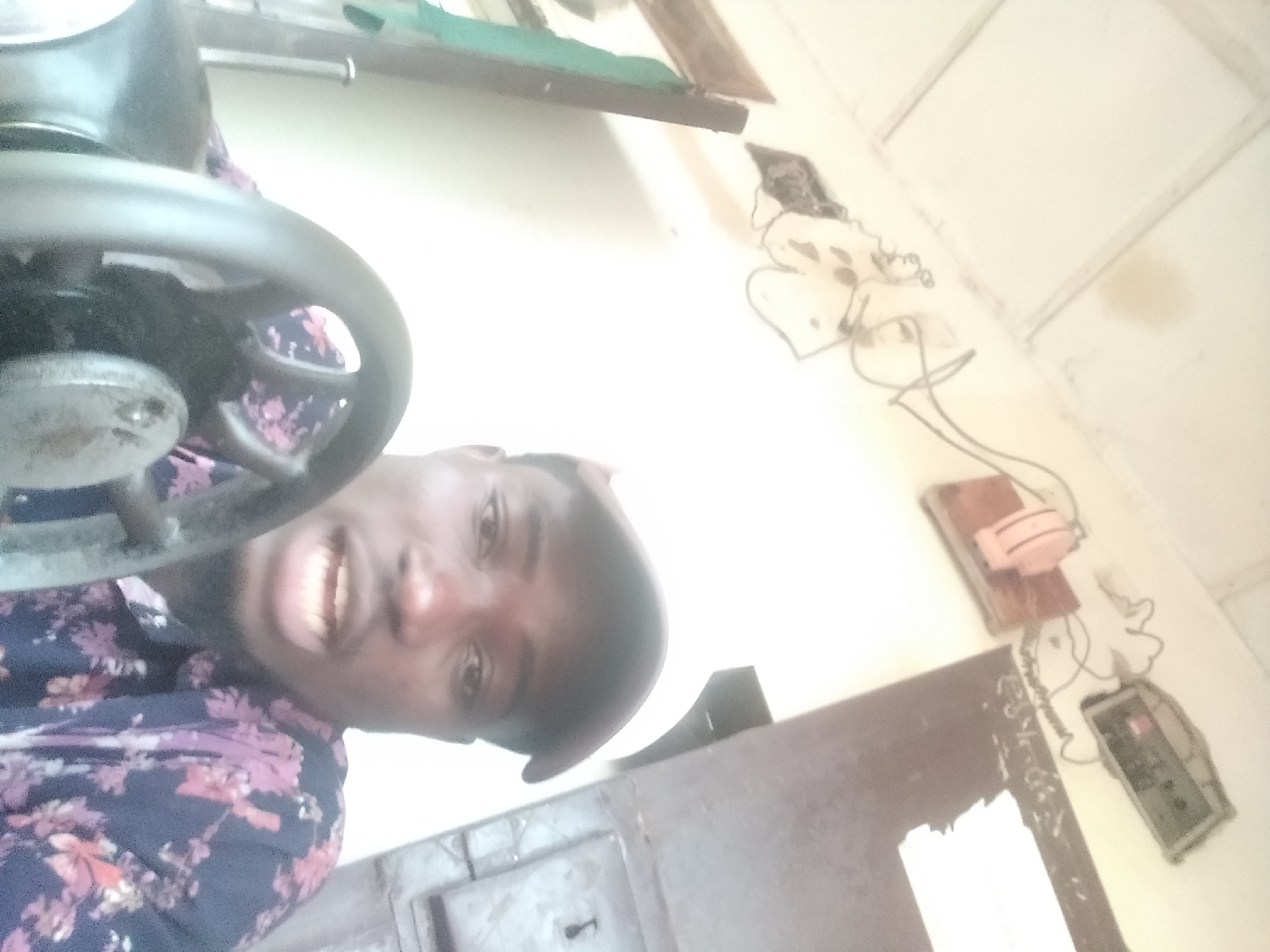 In Jalingo, Nigeria, a young Black male wearing a white baseball cap and a dark blue shirt with a floral pattern is seated in front of a sewing machine. He is looking towards the camera with his mouth slightly open. To the left of the man, a green cloth is hanging from a wooden frame. Above and behind him, wires are strung across the wall, connected to a pink electrical device and a grey electronic box. The scene appears to be indoors, possibly a workshop or residential space, with a white ceiling and a dark wooden door in the background.