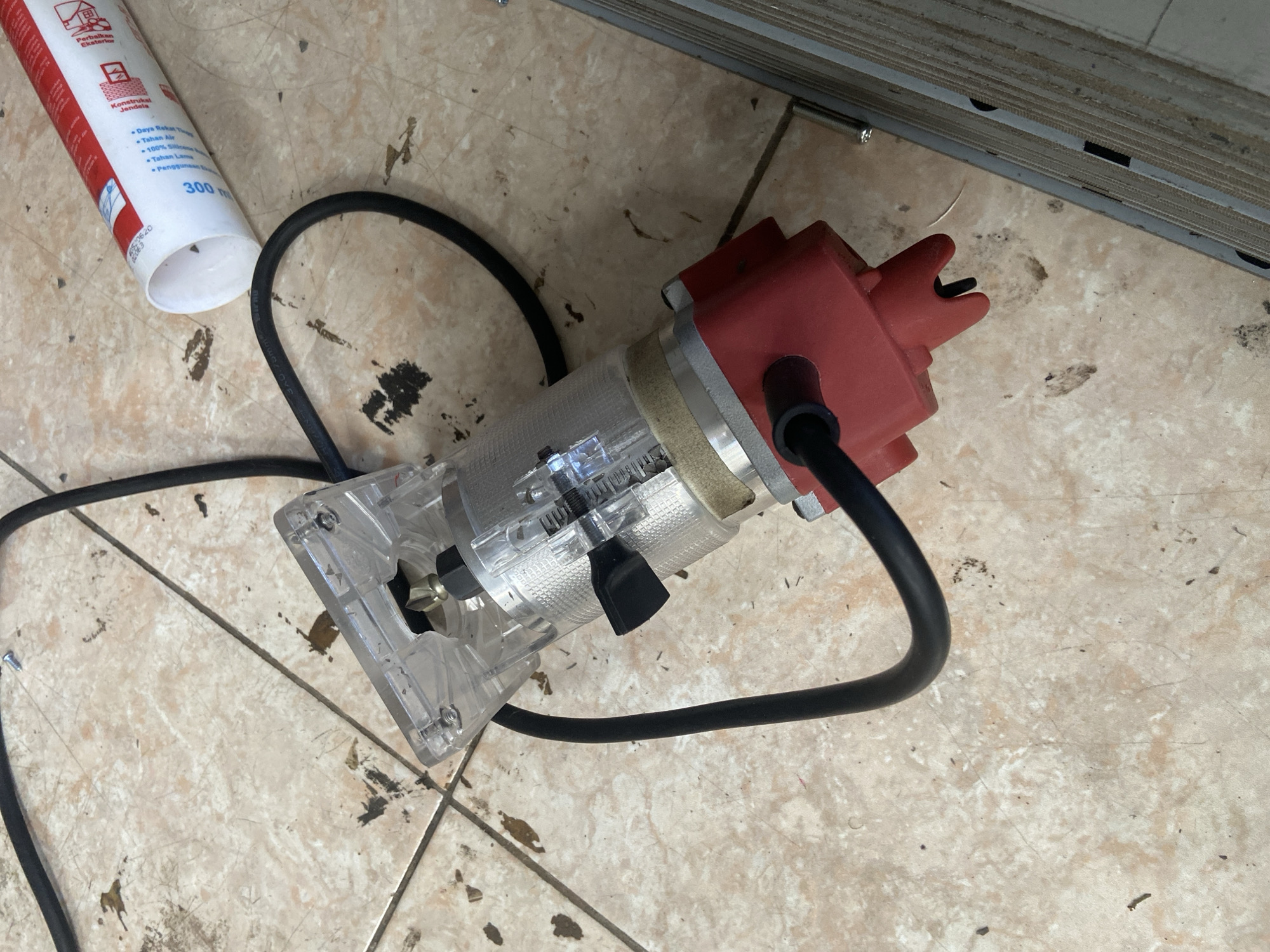 A red and clear plastic rotary tool is lying on a tiled floor with a black power cord extending to the upper left. A white and red cylindrical tube is partially visible to the right of the rotary tool. The tiled floor is a light tan with brown and black debris scattered across its surface. The scene appears to be indoors, possibly a workshop or garage. There are no people or actions depicted in the image. The specific location of Plosokandang, Indonesia is not visually represented in the image.